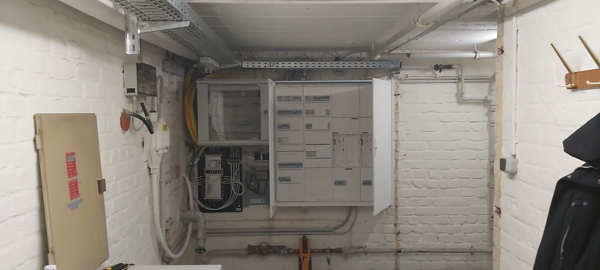 Otwarta skrzynka elektryczna z widocznymi bezpiecznikami i przewodami w pomieszczeniu z pomalowanymi na biało ceglanymi ścianami i instalacją pod sufitem.