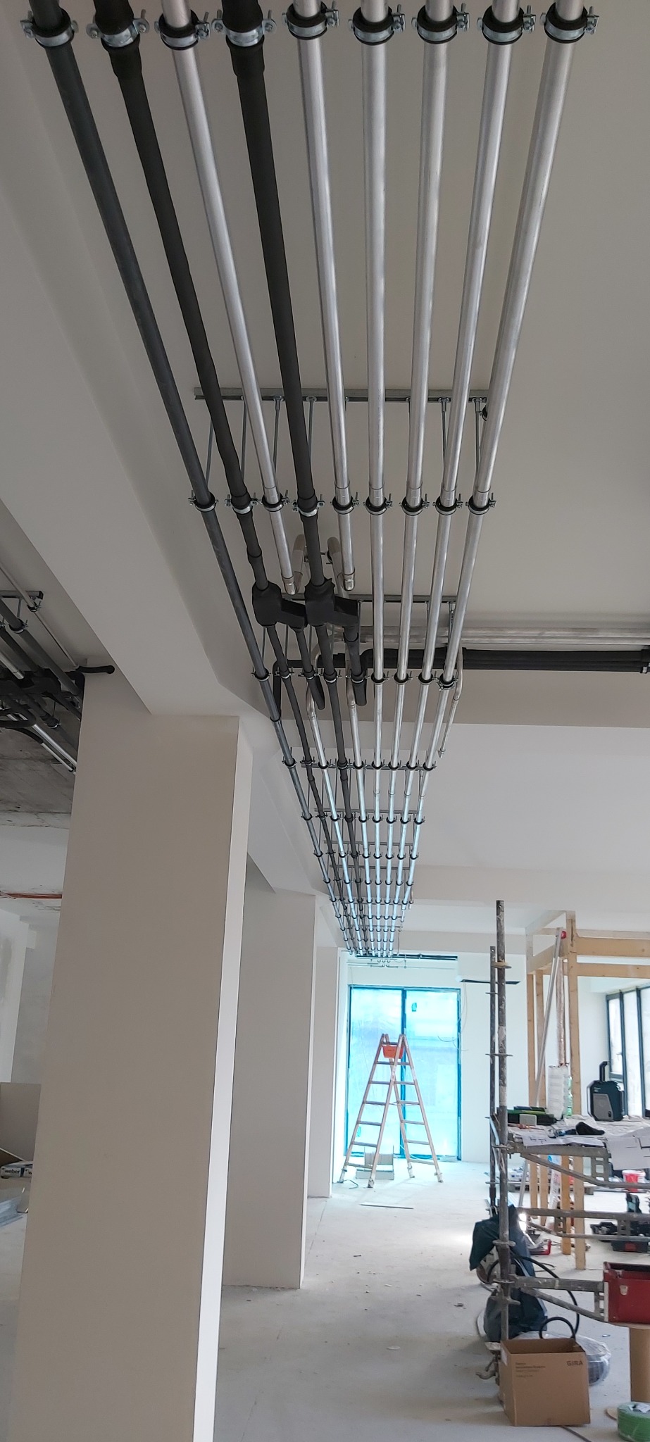 Równoległe rzędy rur instalacyjnych, częściowo w kolorze srebrnym, częściowo czarnym, zamocowane do sufitu za pomocą metalowych uchwytów, widoczne w surowym wnętrzu budynku w trakcie prac...