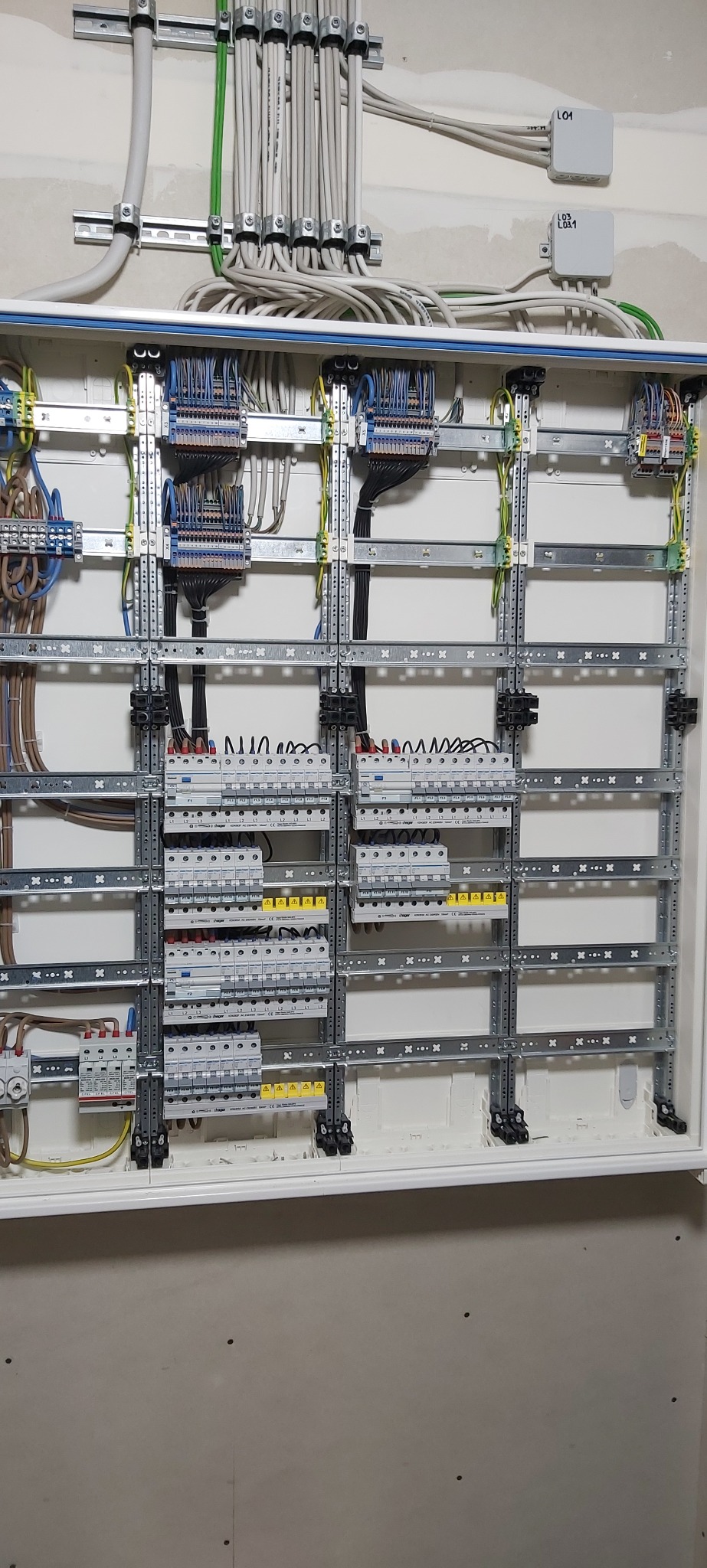 Rozdzielnica elektryczna w trakcie montażu, widoczne kable, listwy montażowe, bezpieczniki i inne komponenty, częściowo okablowana, z białym tłem wewnątrz obudowy.