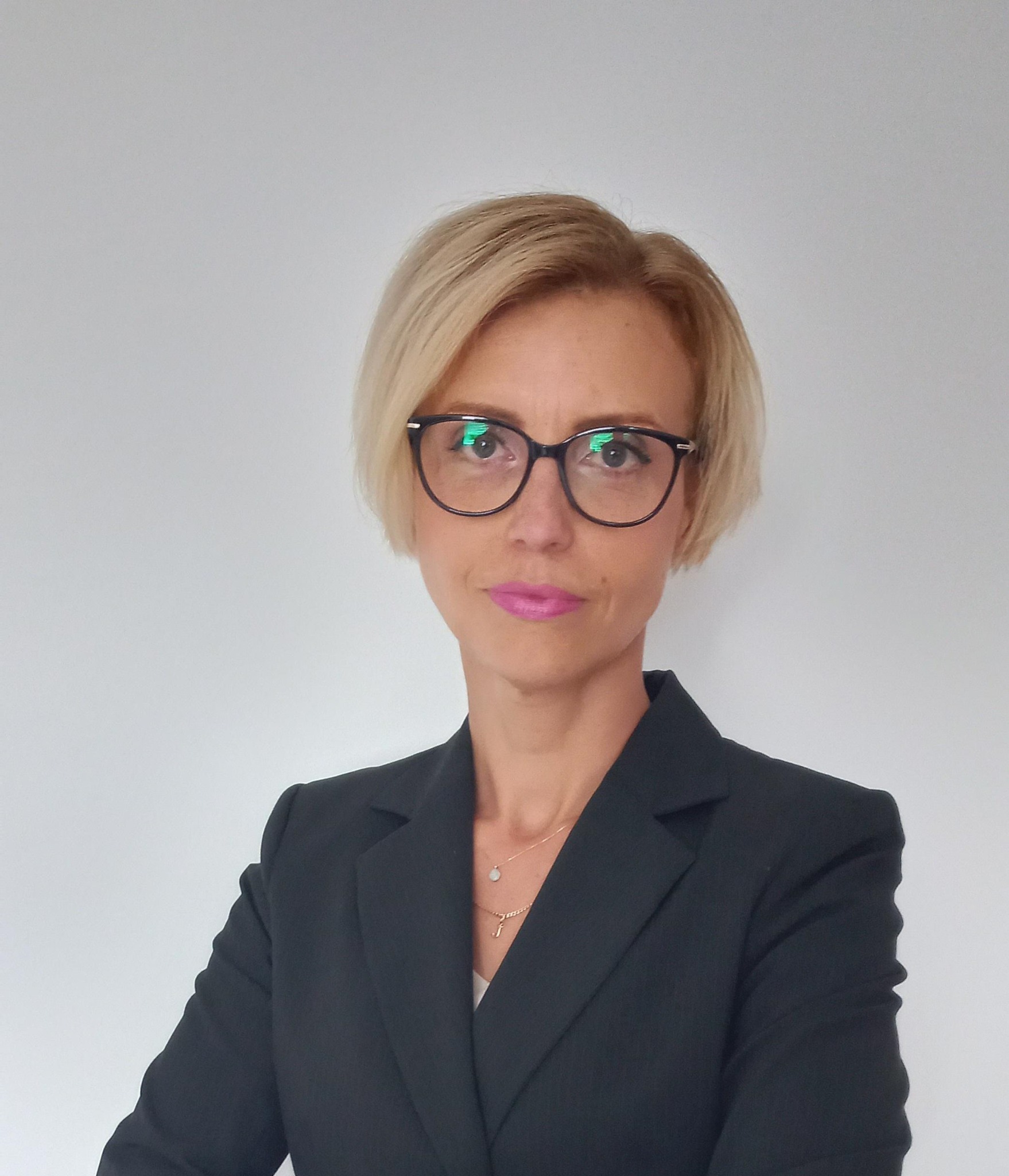 Portret kobiety w czarnym żakiecie i okularach, z krótkimi blond włosami i różową szminką, na jasnym tle.