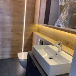 Nowoczesna łazienka z drewnopodobnymi płytkami, podświetlanym lustrem i białą umywalką nablatową. Umywalka i fragment lustra zabezpieczone folią ochronną po montażu.