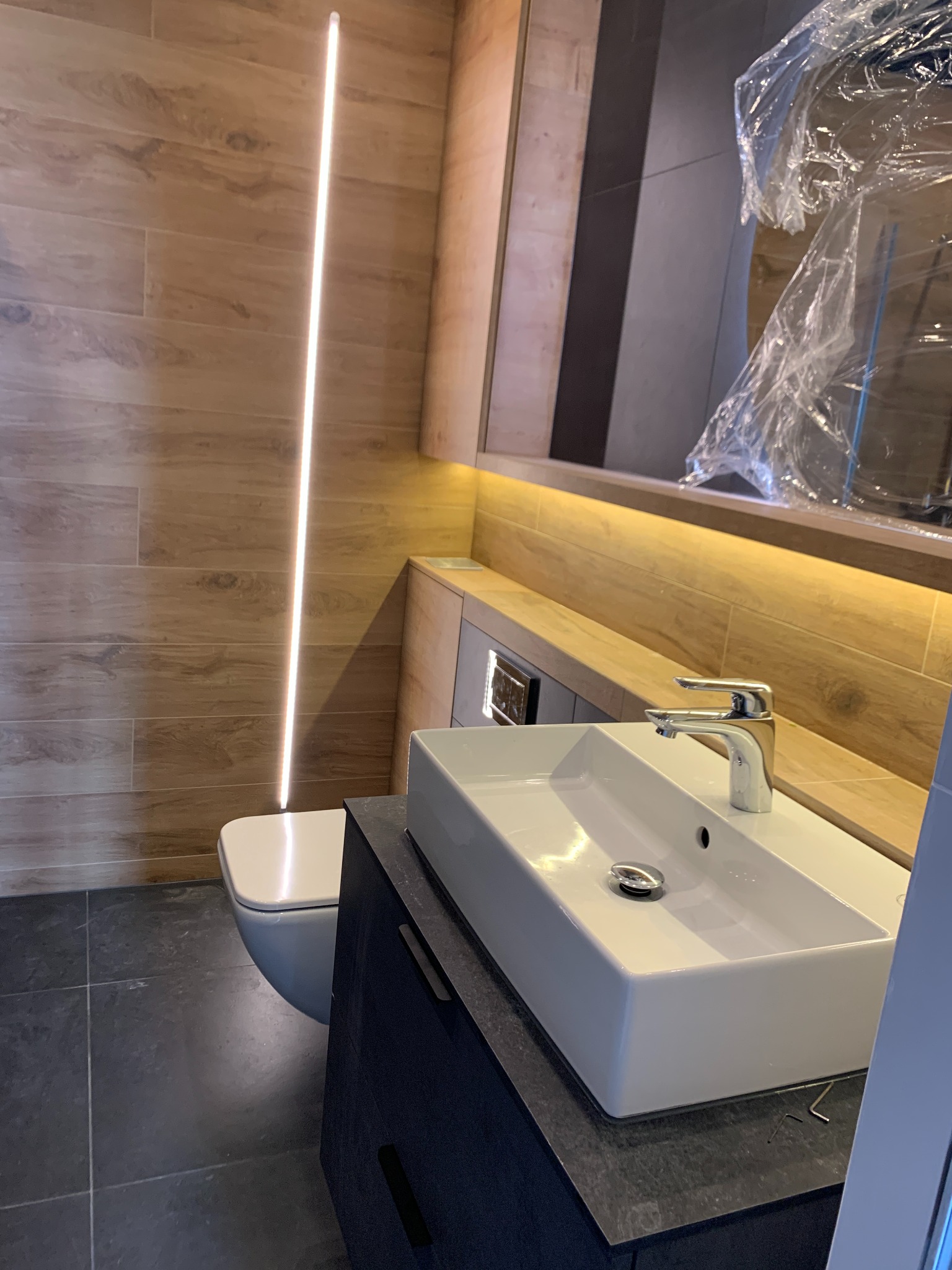 Nowoczesna łazienka z drewnopodobnymi płytkami, podświetlanym lustrem i białą umywalką nablatową. Umywalka i fragment lustra zabezpieczone folią ochronną po montażu.