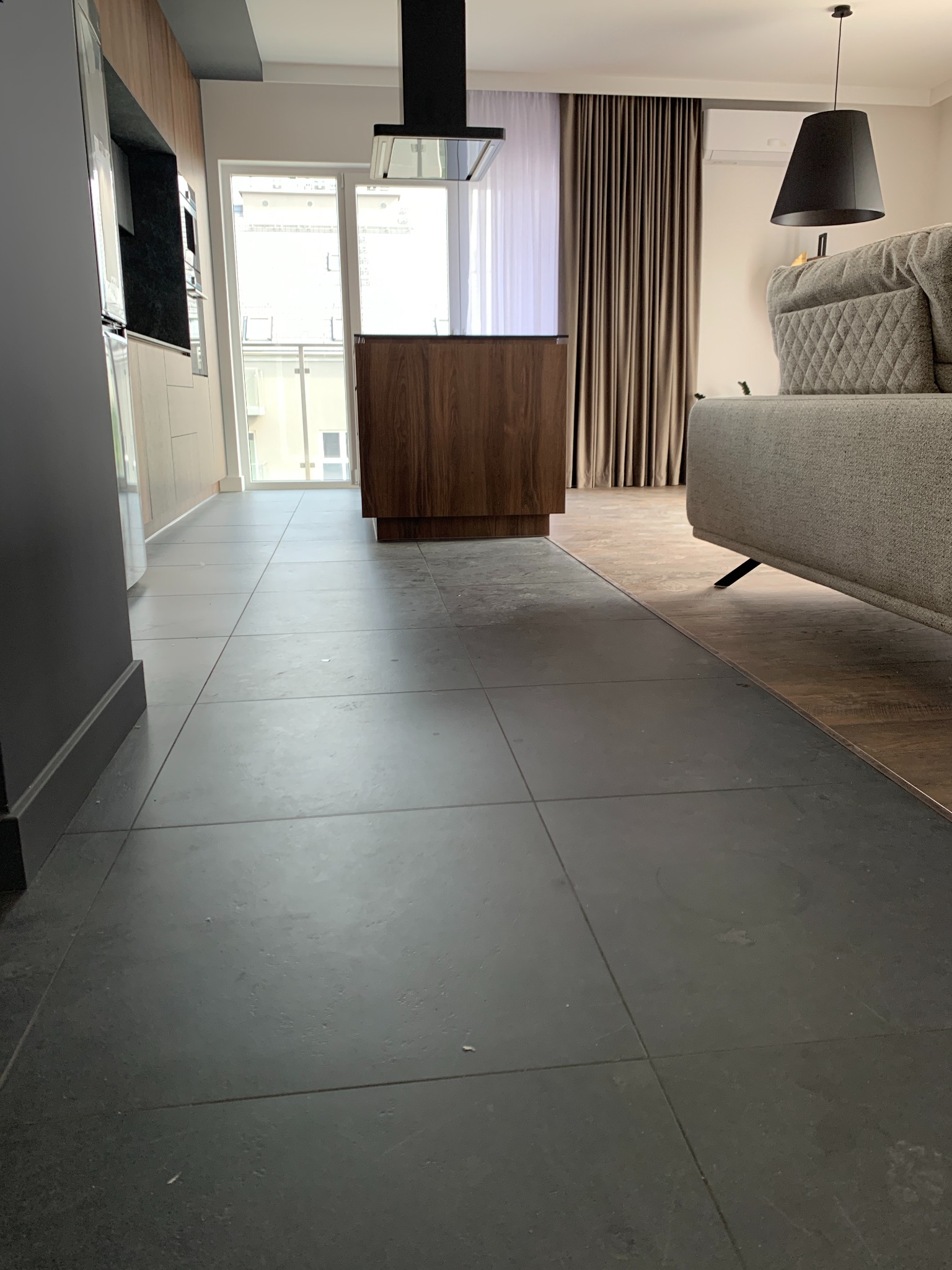 Szare płytki podłogowe, widoczne ślady po remoncie, fragment sofy i elementów kuchennych w tle.