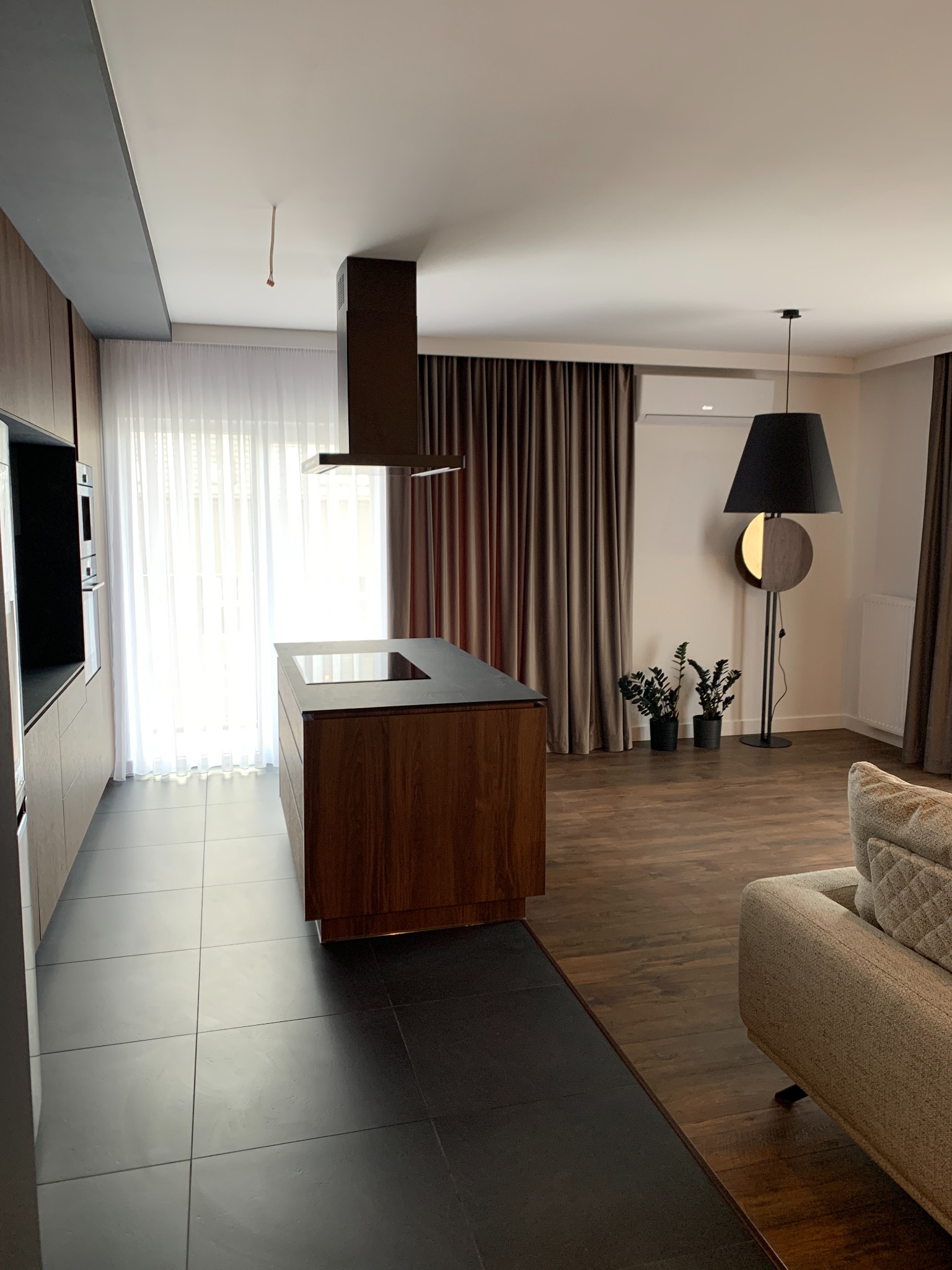 Nowoczesna kuchnia z wyspą kuchenną i ciemnymi płytkami podłogowymi, widok na salon z sofą i lampą podłogową, minimalistyczny design.