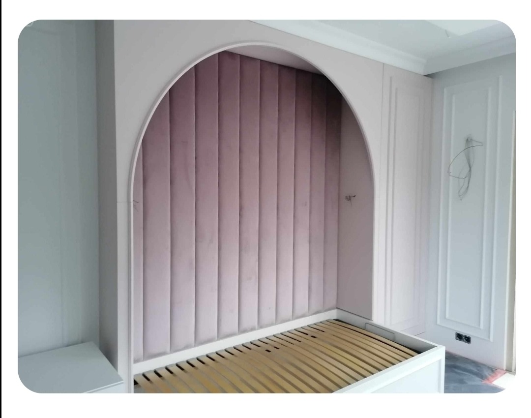 Wnęka łóżka z wezgłowiem tapicerowanym w pionowe pasy w odcieniach pudrowego różu, obudowana łukiem z białego MDF, widoczny stelaż łóżka z listew drewnianych.