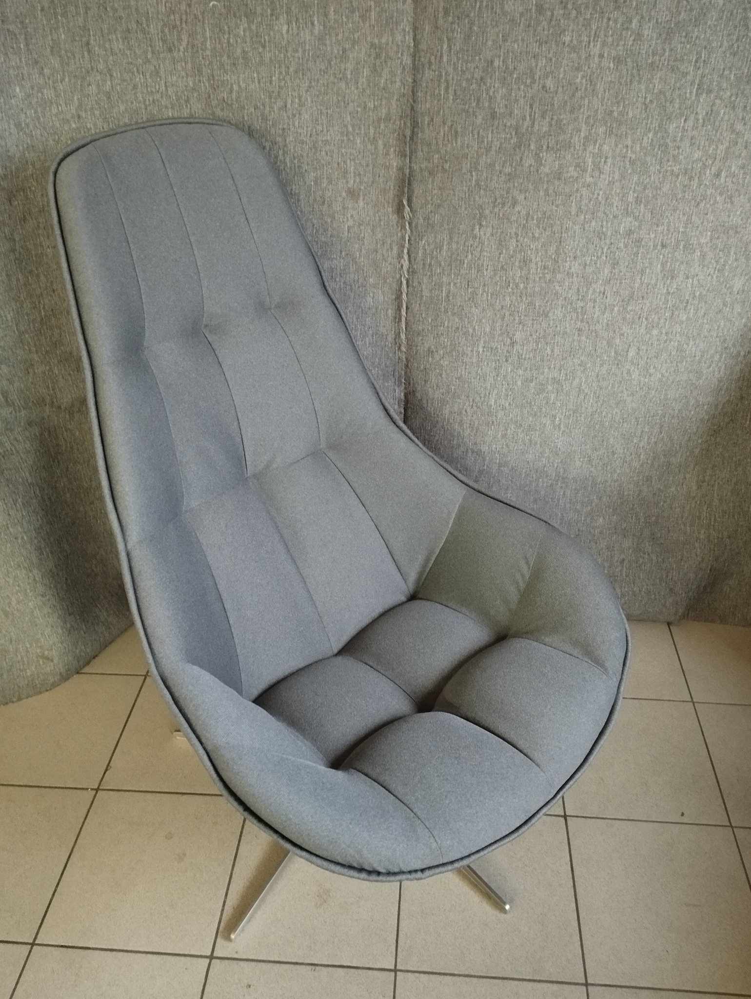 Nowoczesny fotel tapicerowany w odcieniach szarości z pikowaniem, na metalowej podstawie z trzema nogami, ustawiony na płytkach podłogowych.