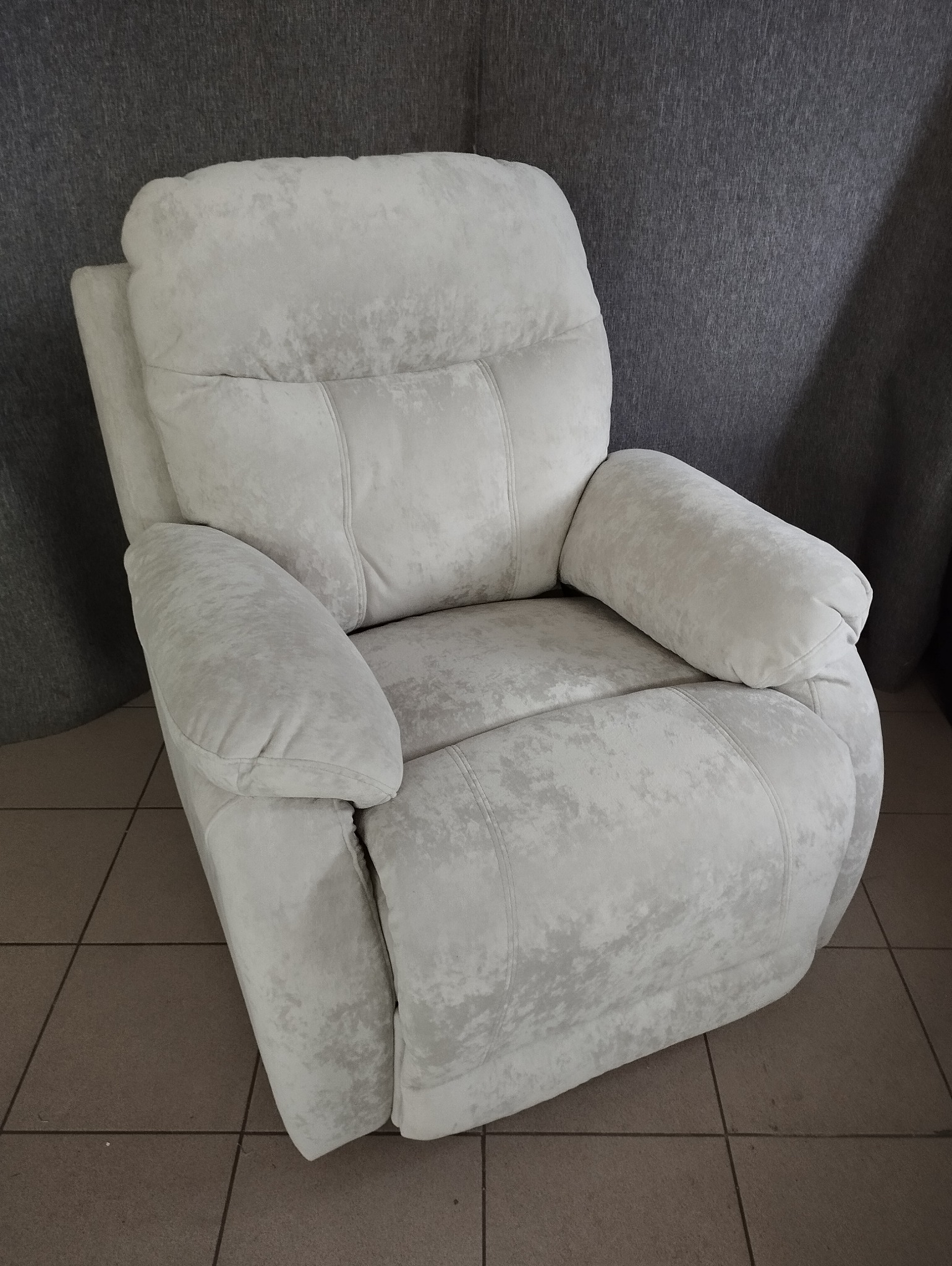 Jasny, tapicerowany fotel relaksacyjny z podłokietnikami, na tle szarej ściany i brązowej podłogi z płytek.