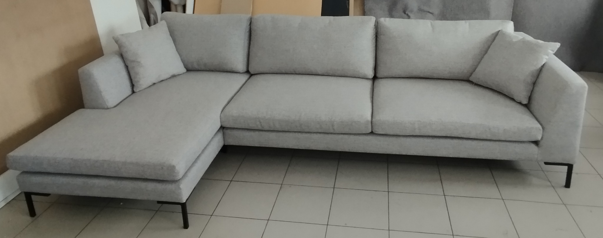 Szara, tapicerowana sofa narożna z luźnymi poduszkami i czarnymi, metalowymi nóżkami, ustawiona na kafelkach podłogowych.