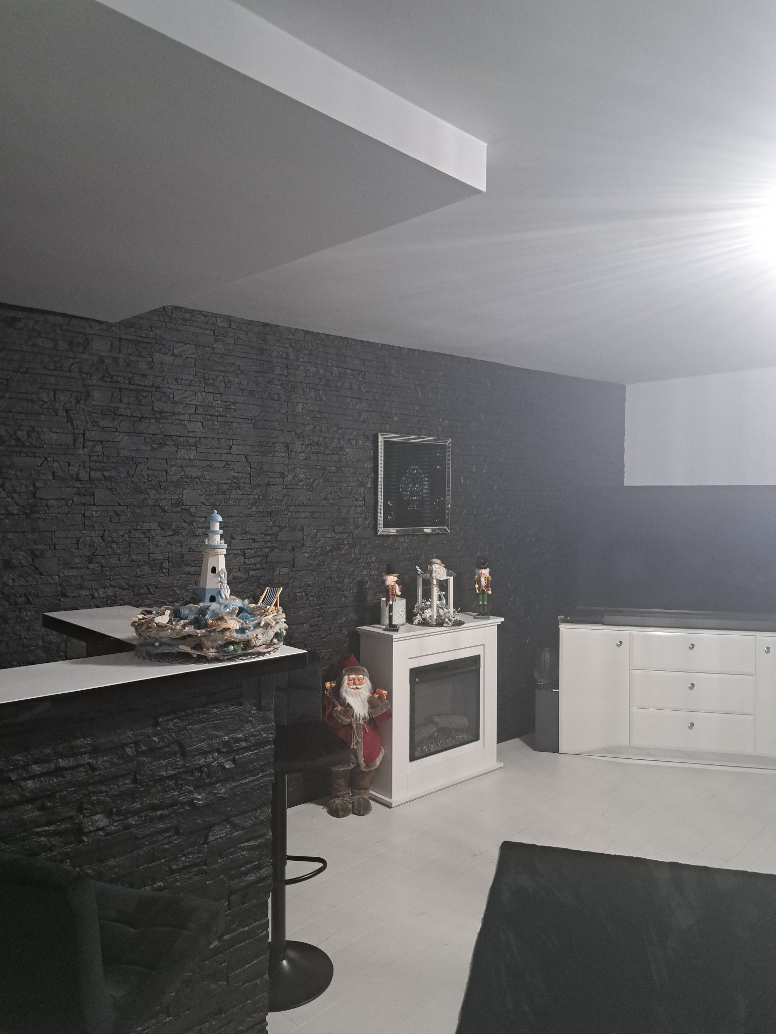 Ciemna ściana z kamienia dekoracyjnego, oświetlona punktowo, z barkiem i dekoracjami świątecznymi, w tle białe meble.