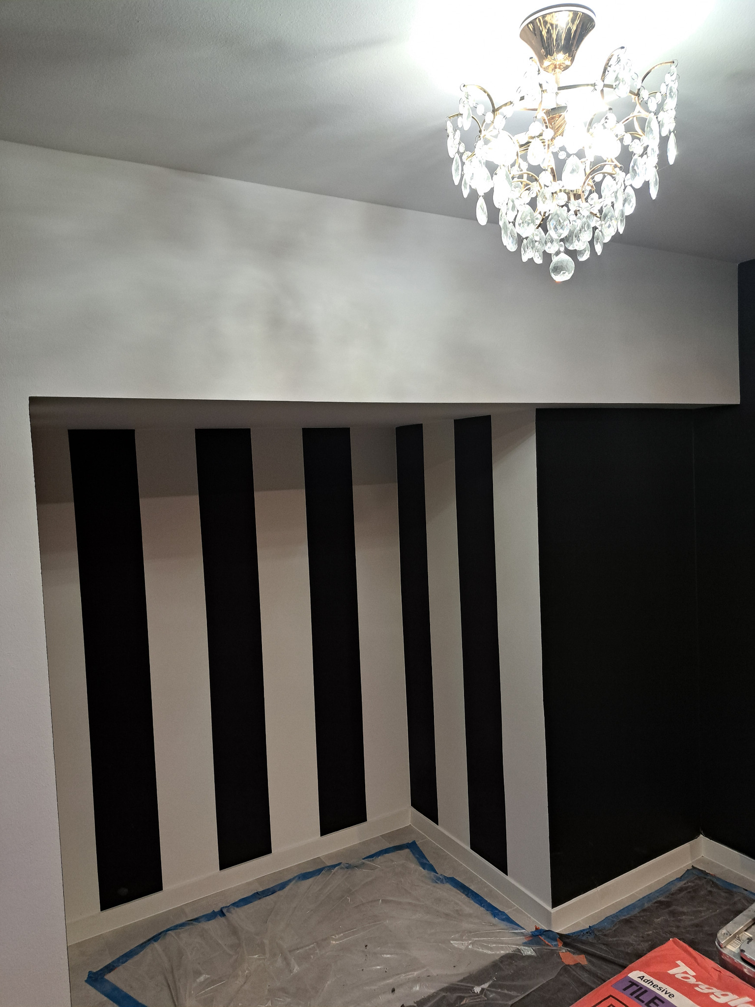 Wnętrze pokoju z pomalowanymi ścianami w czarno-białe pasy, zabezpieczona podłoga folią, widoczny żyrandol kryształowy, częściowo pomalowana ściana na czarno.