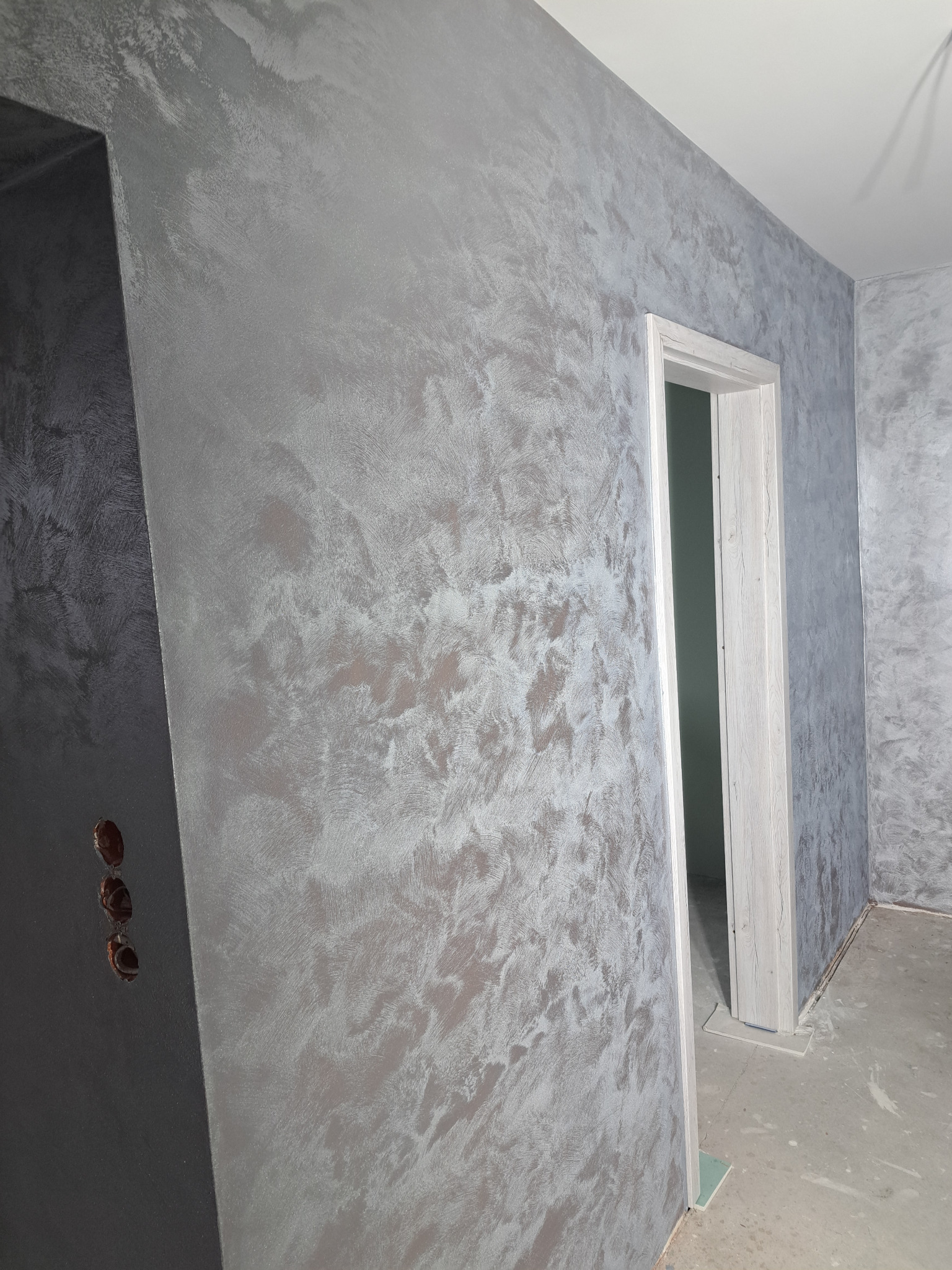 Ściana pokryta dekoracyjną, metaliczną farbą w odcieniach szarości i srebra, widoczna futryna drzwi i niezamaskowane otwory pod gniazdka elektryczne.