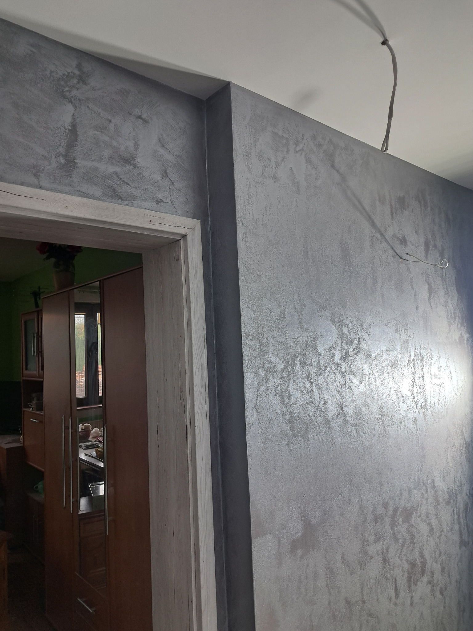 Ściana z efektem dekoracyjnym w odcieniach szarości, widoczna obok futryny drzwiowej z drewnopodobnym wykończeniem i wystającym przewodem elektrycznym z sufitu.