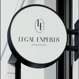 Kancelaria Adwokacka adwokat Izabela Raszeja - Elegancki szyld okrągły z napisem 'Legal Experts Adwokaci' i logo LE, zamocowany na białej fasadzie budynku. Minimalistyczny design, czarne elementy na jasnym tle.