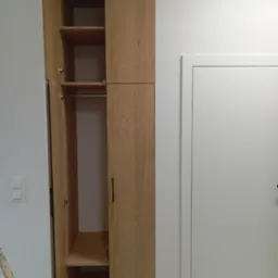 Jasna szafa na wymiar z otwartymi drzwiami, widoczne półki i drążek na ubrania. Obok białe drzwi i włącznik światła na ścianie. Minimalistyczny design.