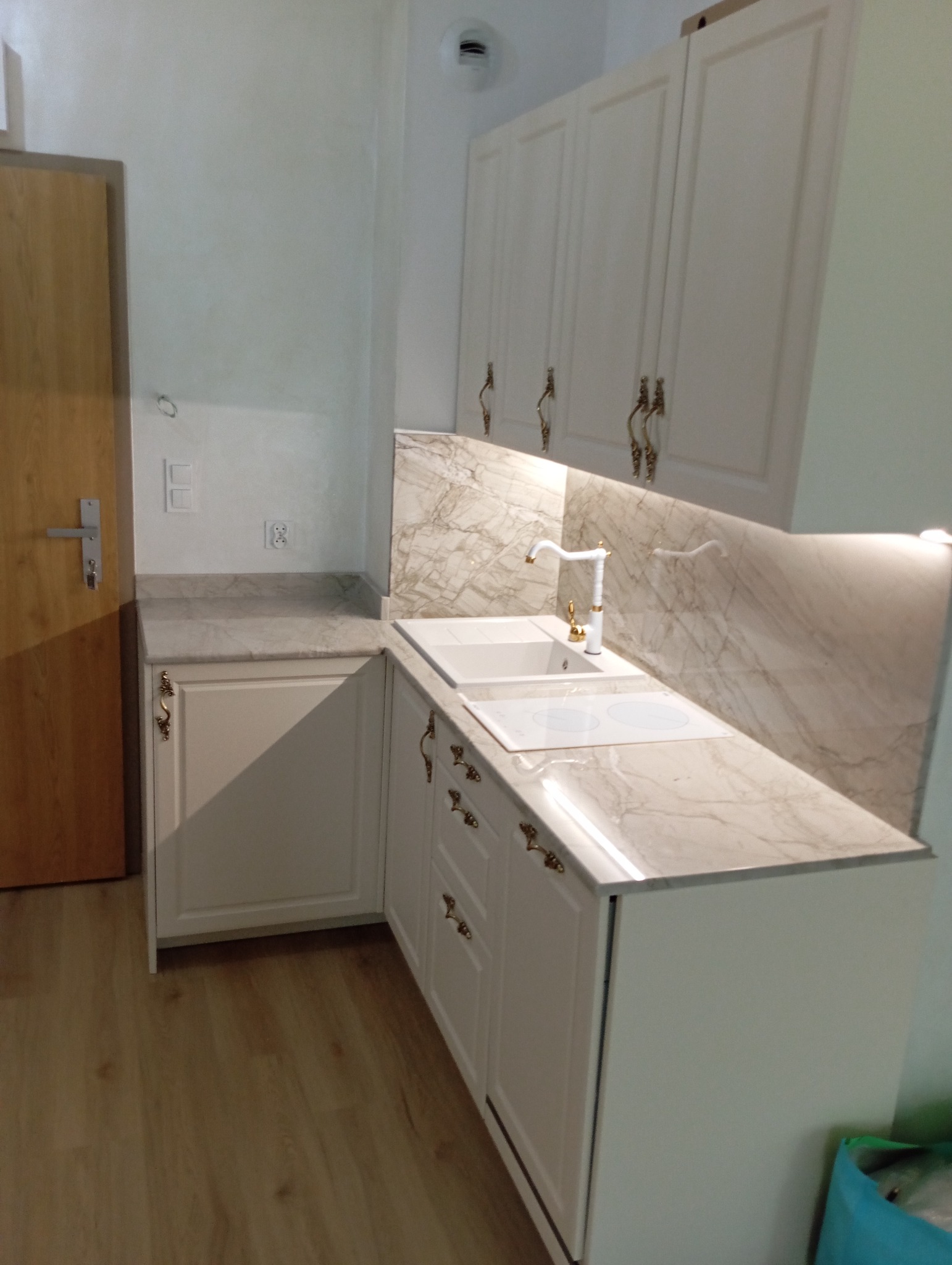 Jasna kuchnia z marmurowym blatem i złotymi uchwytami. Widoczne szafki wiszące i dolne, zlew oraz płyta grzewcza. Uwagę przykuwa bateria w stylu retro.