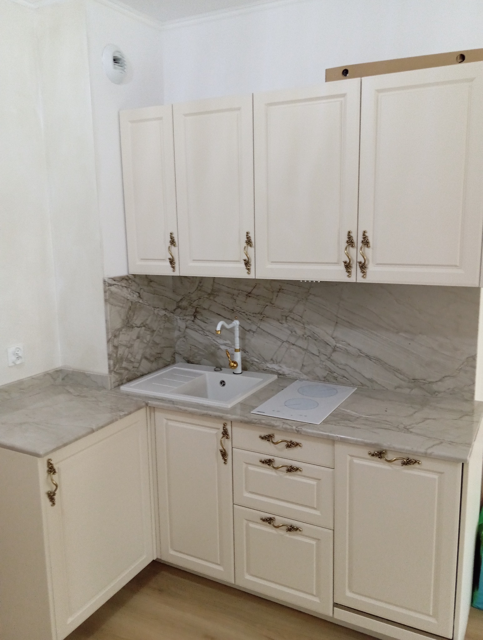 Jasna zabudowa kuchenna z marmurowym blatem i ozdobnymi uchwytami. Widoczny zlew, płyta indukcyjna i szafki górne. Klasyczny styl.