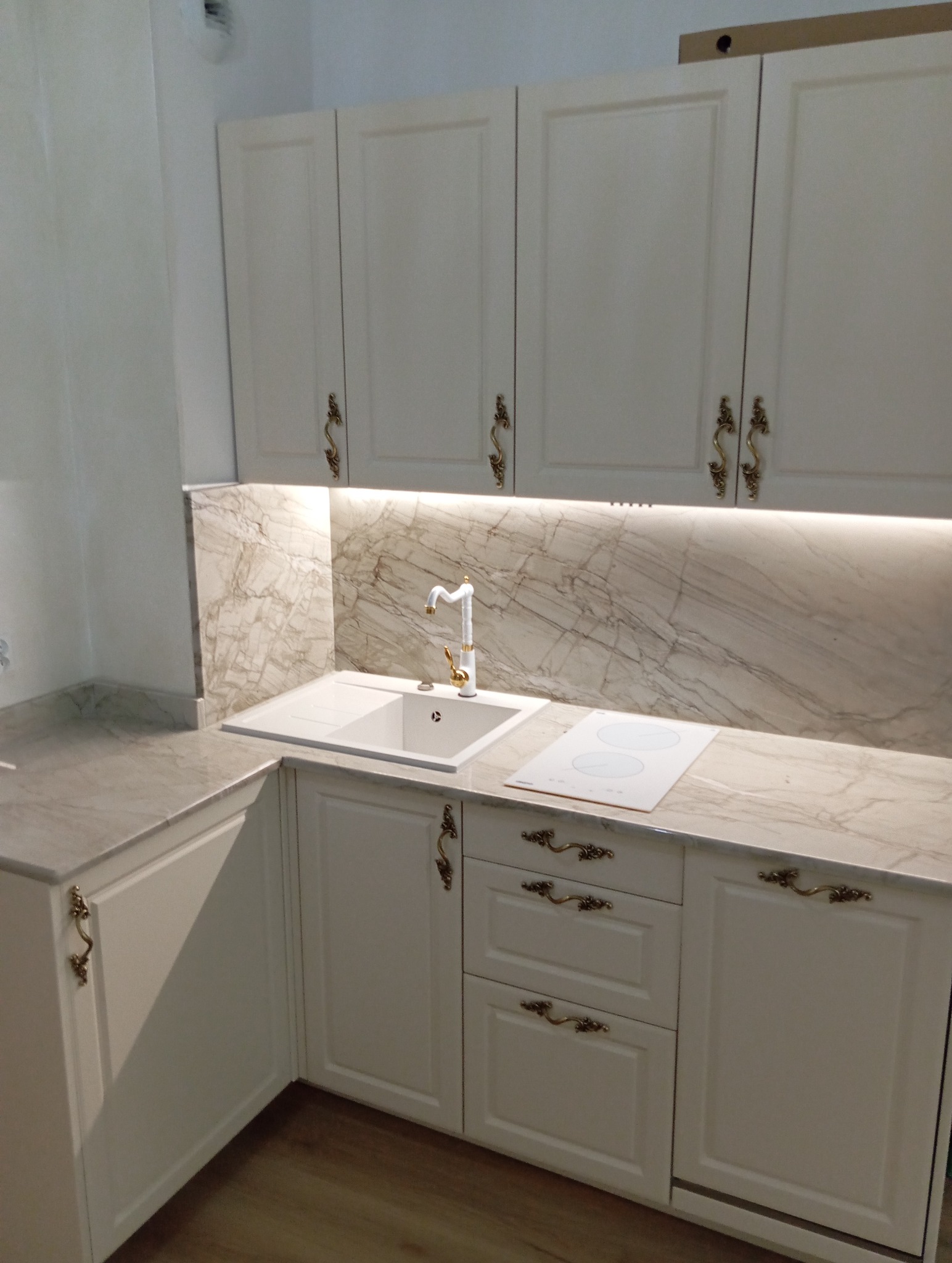 Jasna kuchnia z białymi frontami, marmurowym blatem i złotymi uchwytami. Uwagę zwraca podświetlenie nad blatem i nietypowa bateria.