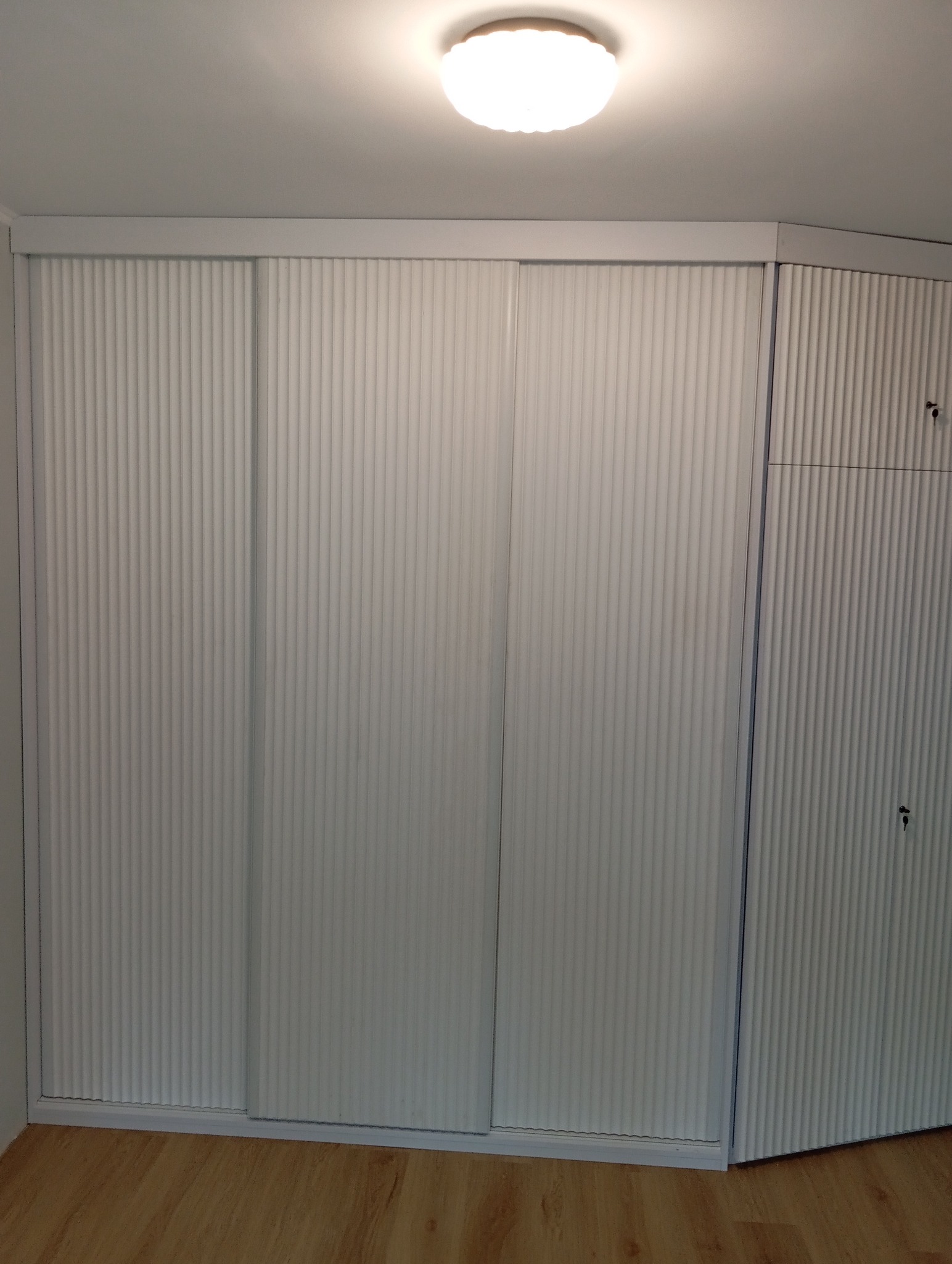 Biała szafa wnękowa z przesuwnymi drzwiami z charakterystycznym żłobkowaniem, dopasowana do skosu pomieszczenia, z widocznym oświetleniem sufitowym.