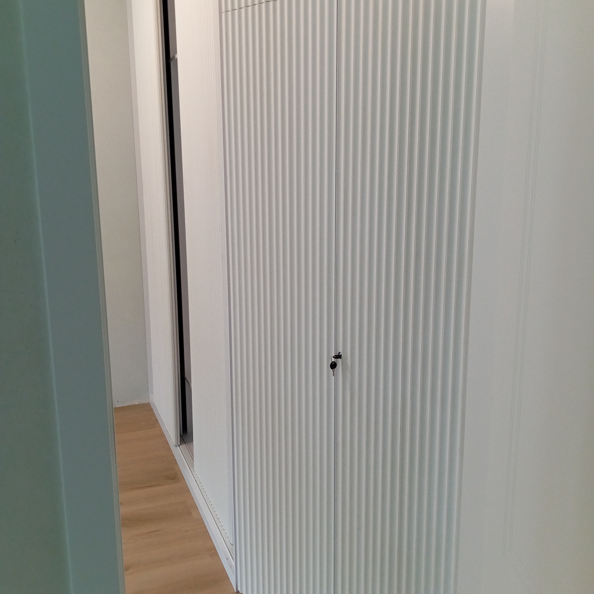 Biała szafa wnękowa z ryflowanymi drzwiami, zamykana na klucz, widoczna w korytarzu z panelami podłogowymi. Minimalistyczny design.