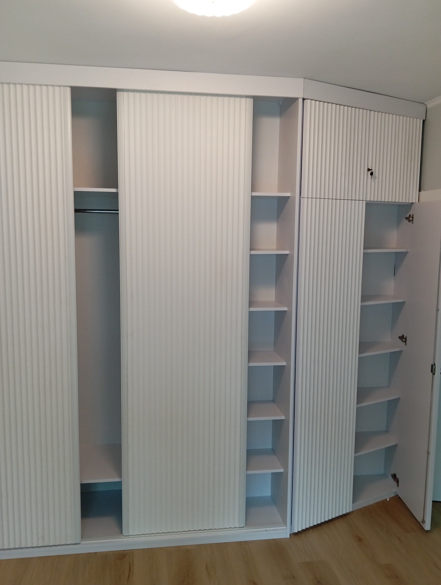 Biała szafa wnękowa z przesuwnymi drzwiami i ryflowanymi frontami. Widoczne półki i drążek na ubrania. Szafa dopasowana do skosu sufitu.