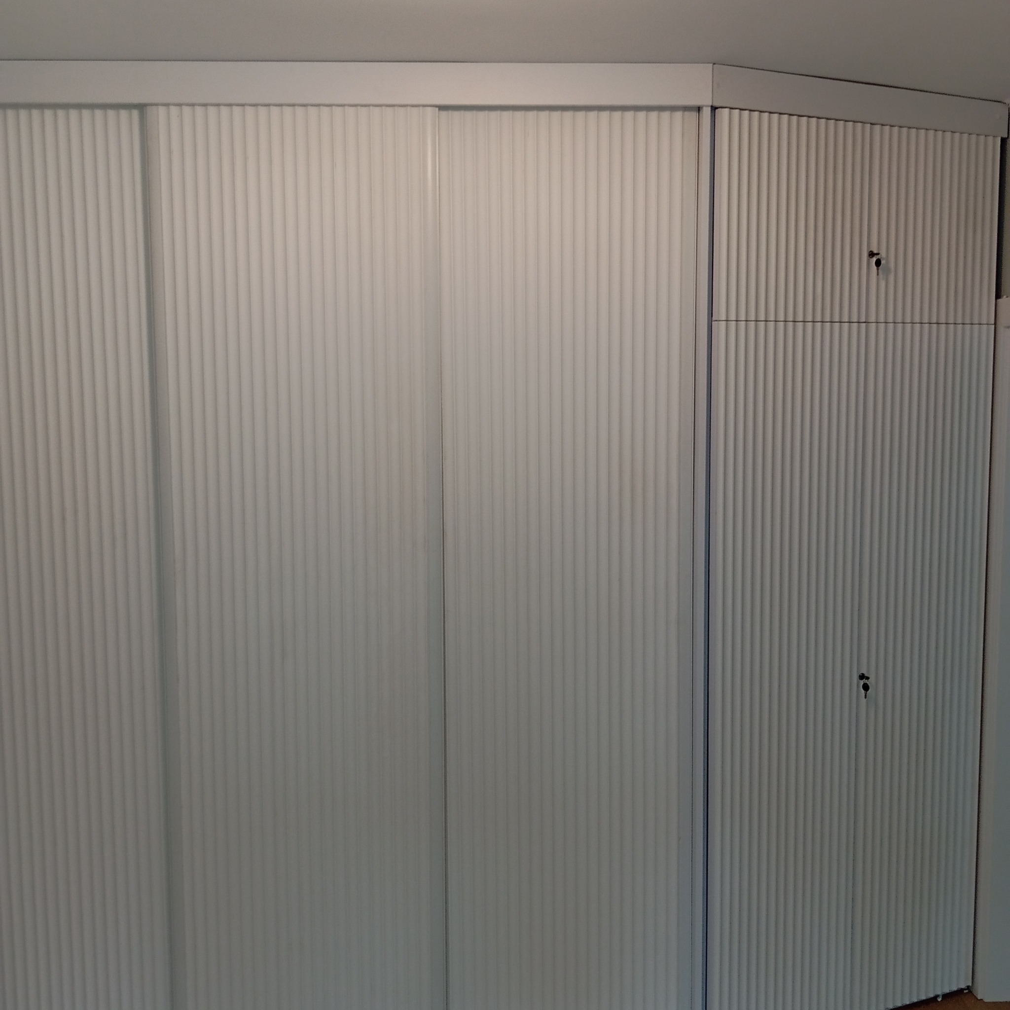 Biała szafa wnękowa z przesuwnymi drzwiami i ryflowanym wykończeniem, dopasowana do narożnika pomieszczenia, z widocznymi zamkami w górnej części.