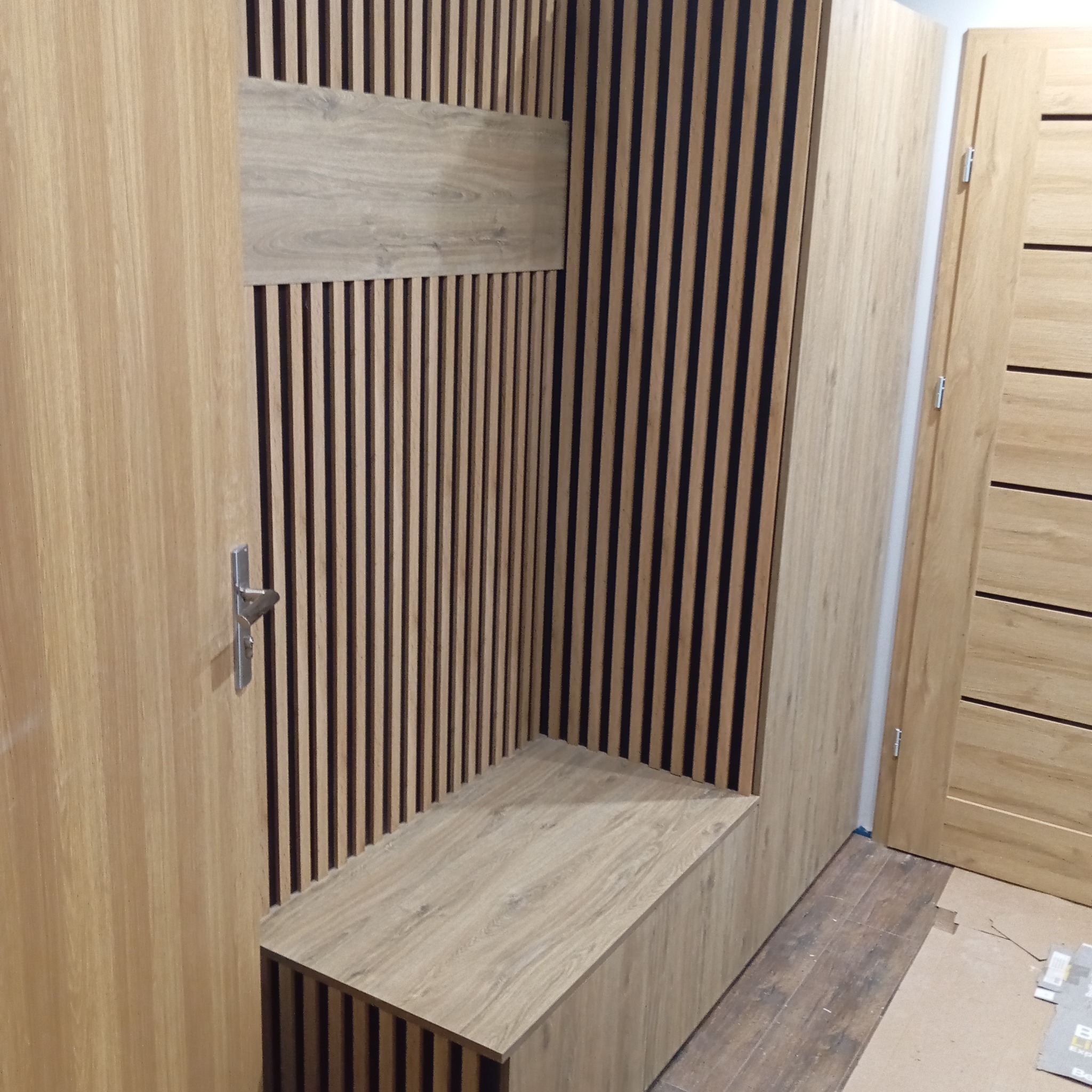 Zabudowa wnęki z siedziskiem i panelami ściennymi w odcieniach drewna. Widoczne drzwi z drewnianą okleiną i metalową klamką. Nowoczesny design.