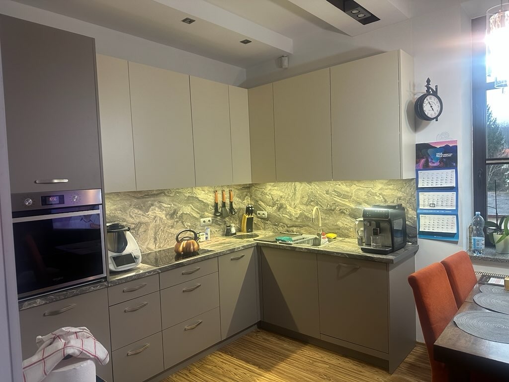 Jasna kuchnia z szarymi frontami i blatem imitującym marmur. Na blacie ekspres do kawy, czajnik i robot kuchenny. Nad blatem wisząca szafka i reling z akcesoriami.