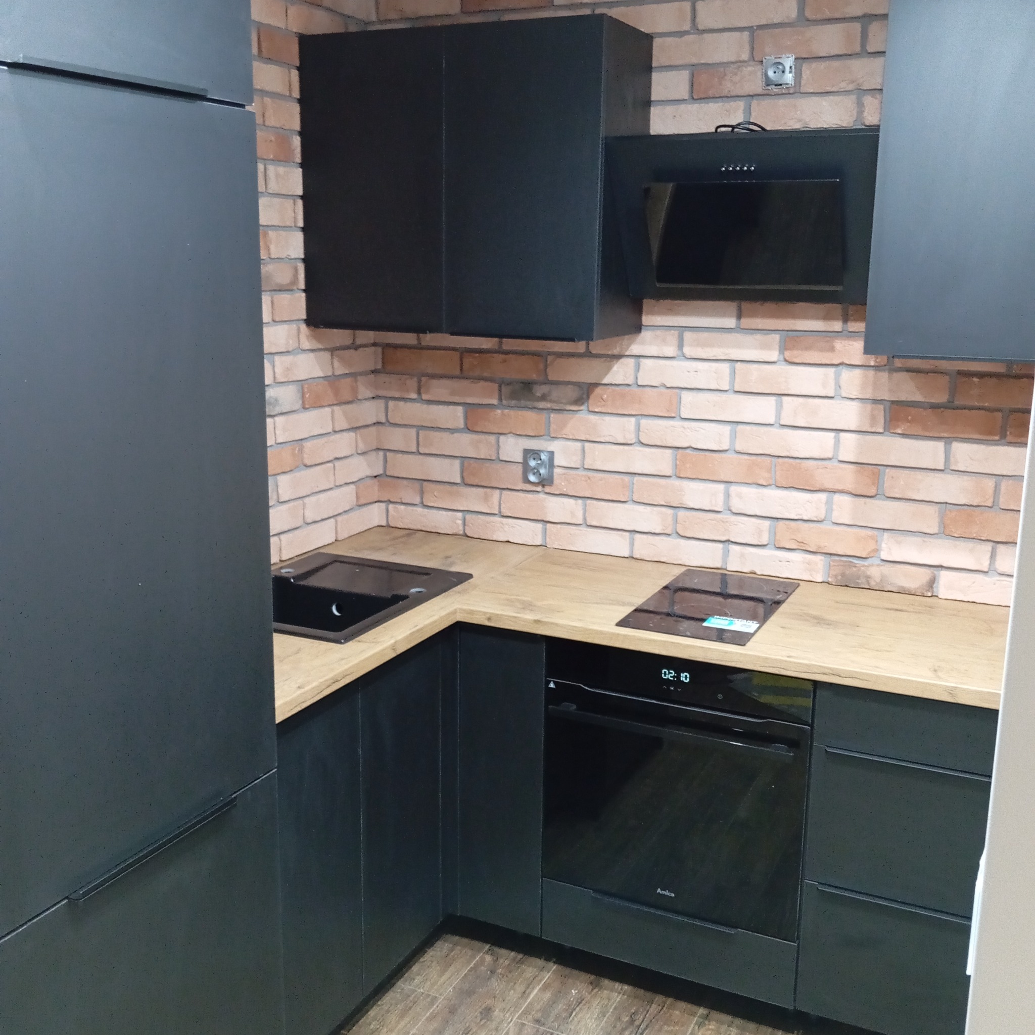 Nowoczesna, czarna kuchnia z blatem imitującym drewno i ceglaną ścianą. Widoczny zlew, płyta indukcyjna i piekarnik. Minimalistyczny design, oświetlenie punktowe.