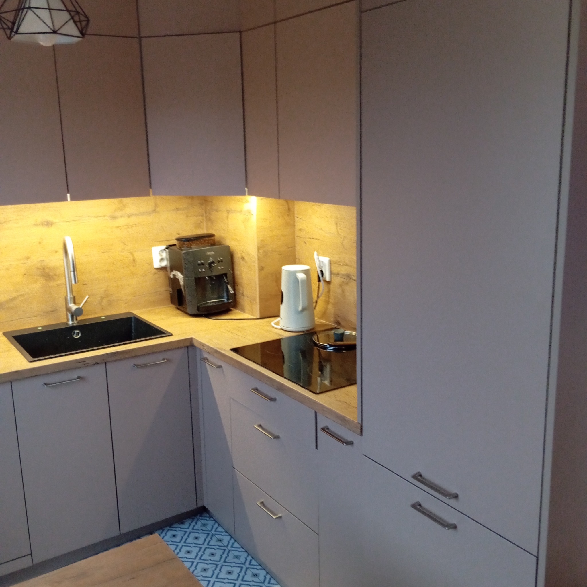 Szara kuchnia z drewnianym blatem, czarnym zlewem i płytą indukcyjną. Widoczne urządzenia: ekspres do kawy i czajnik. Podłoga z wzorem geometrycznym.