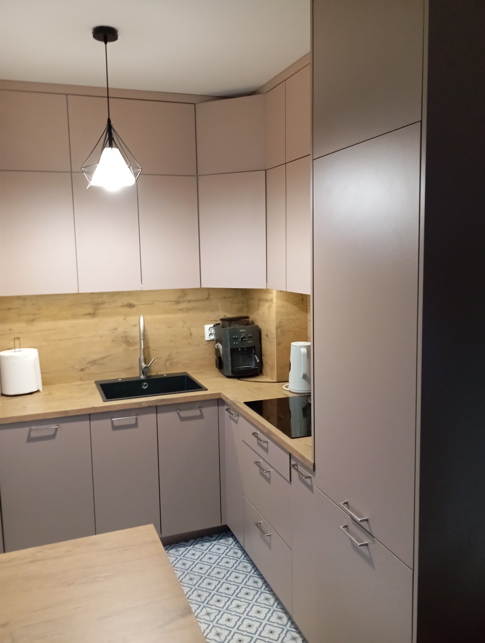 Jasna kuchnia w kształcie litery L z drewnianym blatem i wzorzystą podłogą. Nad blatem wisi nowoczesna lampa z geometrycznym kloszem.