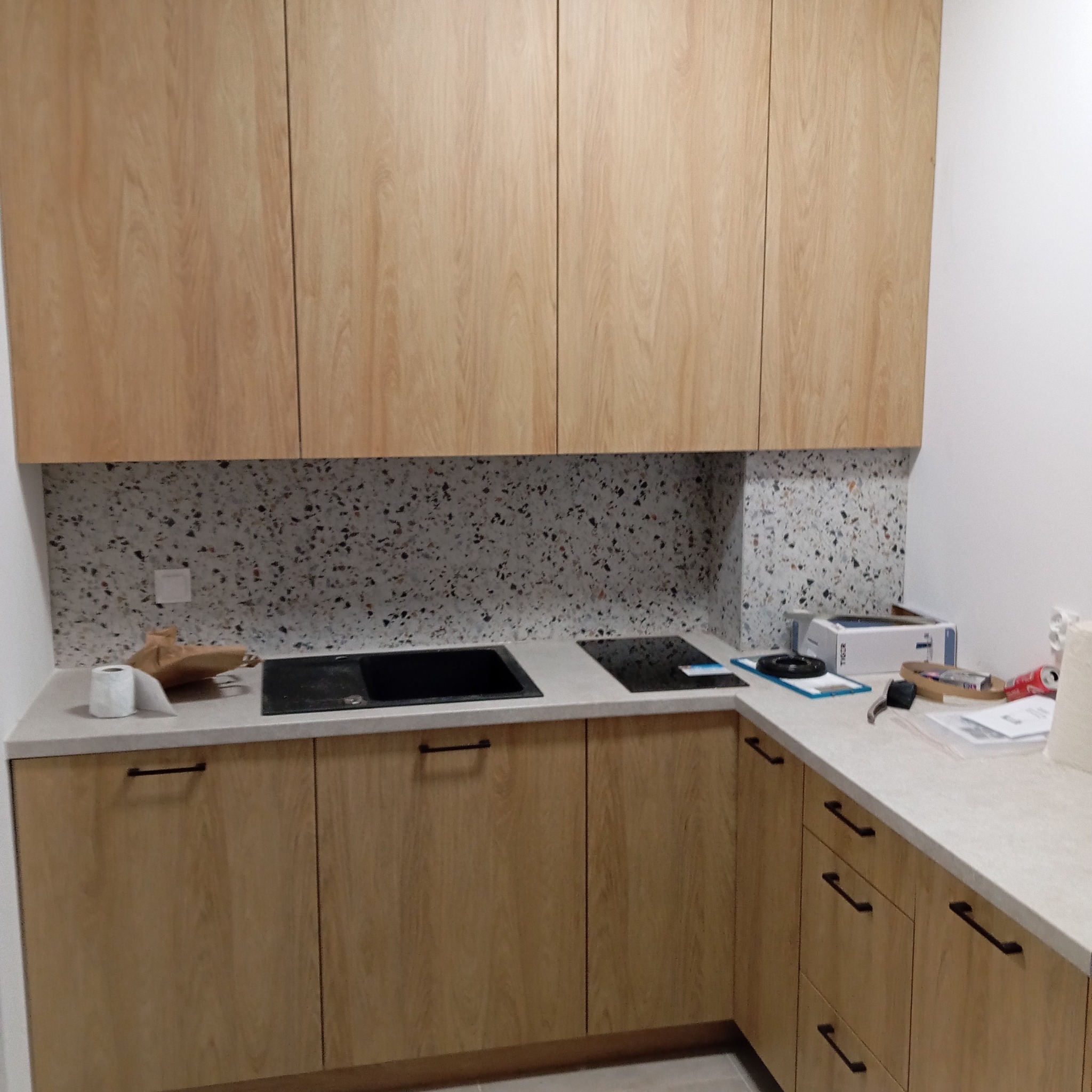 Jasna kuchnia z drewnianymi frontami i blatem z drobnym wzorem. Na blacie zlewozmywak, płyta indukcyjna i akcesoria. Ściana wykończona płytkami typu lastryko.