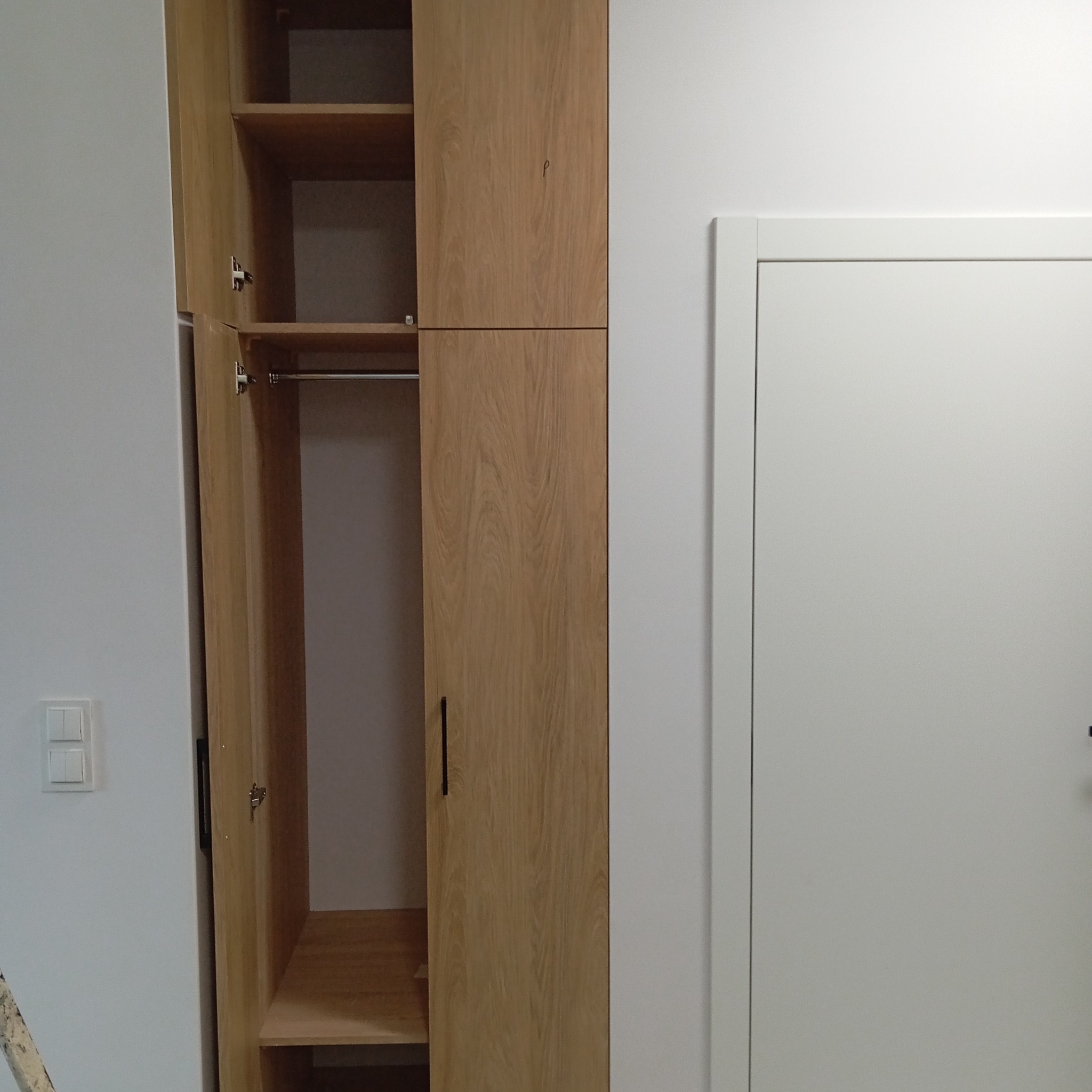 Jasna szafa na wymiar z otwartymi drzwiami, widoczne półki i drążek na ubrania. Obok białe drzwi i włącznik światła na ścianie. Minimalistyczny design.