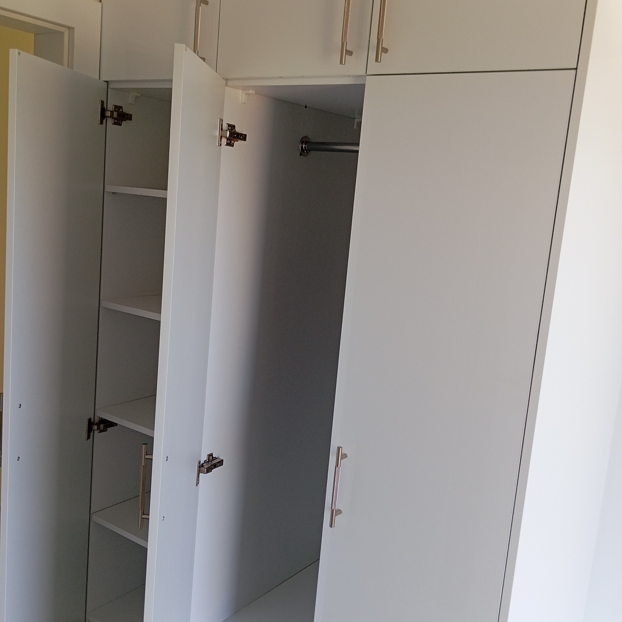 Biała szafa z otwartymi drzwiami, widoczne półki i drążek na ubrania. Minimalistyczny design, metalowe uchwyty. Jasne wnętrze szafy.