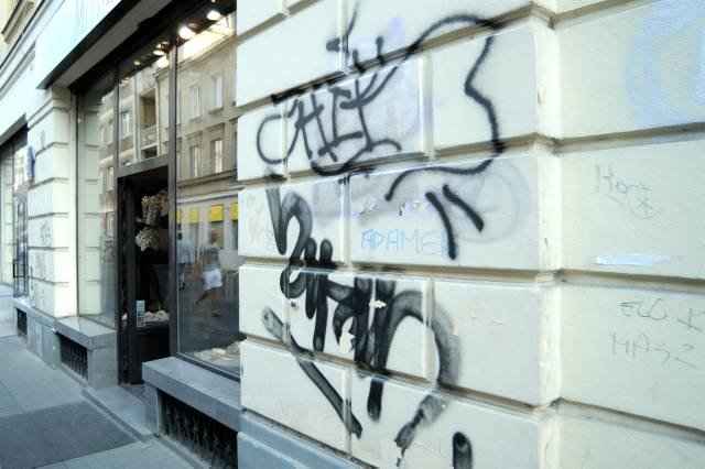 Ściana budynku pokryta graffiti w czarnym kolorze, widoczne napisy i abstrakcyjne wzory, fragment ulicy z wejściem do sklepu w tle.