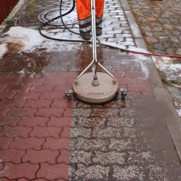 Markowe mycie - Mycie czerwonej kostki brukowej za pomocą myjki ciśnieniowej z okrągłą dyszą; widoczna wyraźna różnica między czystą a brudną powierzchnią, pracownik w pomarańczowym kombinezonie i pachołek...