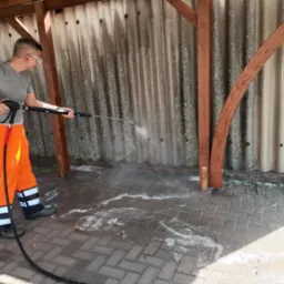 Pracownik w okularach i pomarańczowych spodniach roboczych czyści elewację z blachy falistej myjką ciśnieniową, woda spływa po kostce brukowej.