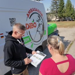 Markowe mycie - Mężczyzna w czarnej bluzie prezentuje kobiecie folder reklamowy na tle firmowego samochodu z logo Dibo i myjką ciśnieniową.