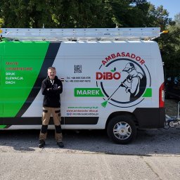 Markowe mycie - Mężczyzna w okularach, ubrany w czarną bluzę i beżowe spodnie robocze, stoi na tle firmowego, zielono-białego busa z napisem 'Mycie ciśnieniowe BRUK Elewacja Dach' oraz logotypem firmy i przyczepą...