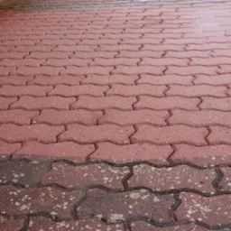 Czerwona kostka brukowa w kształcie zygzaka, częściowo umyta, z widoczną różnicą w kolorze i czystości, z rynną spustową w lewym górnym rogu.