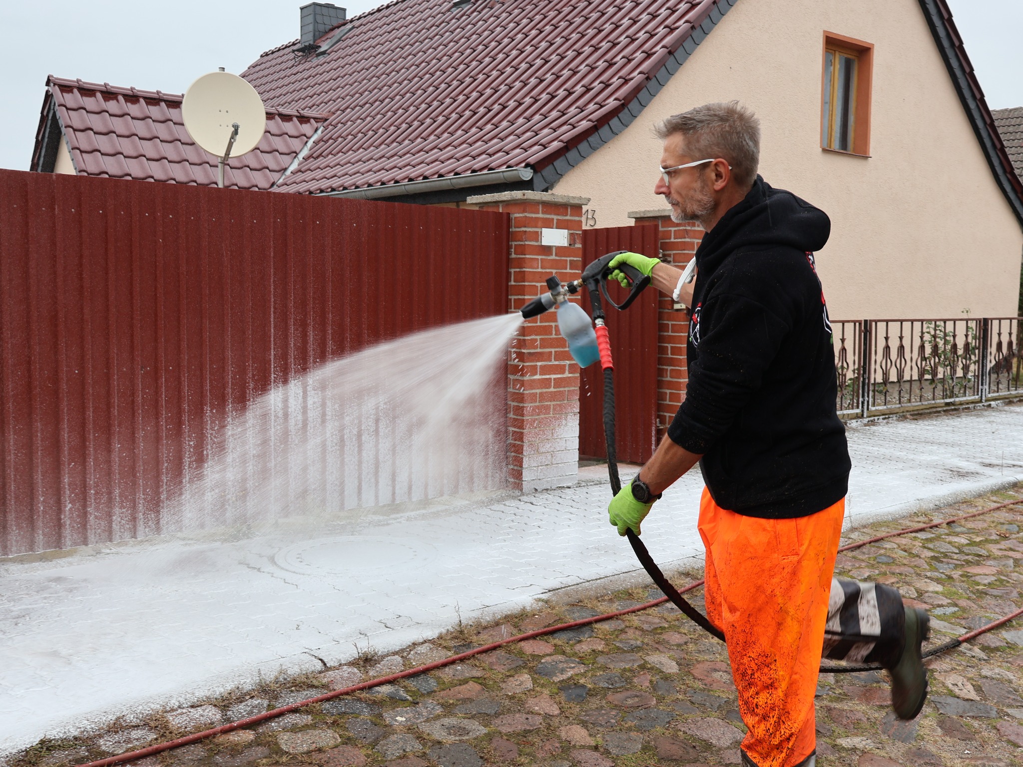 Mężczyzna w pomarańczowych spodniach i czarnej bluzie czyści chodnik z kostki brukowej za pomocą myjki ciśnieniowej, widoczny płot i dom w tle.