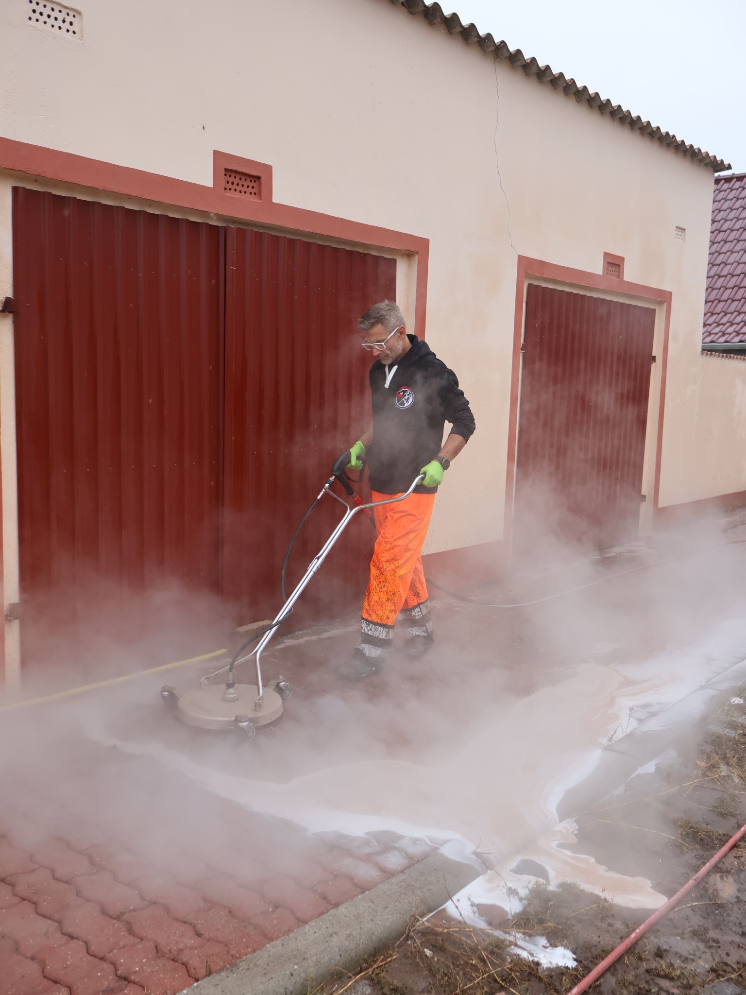 Pracownik w kombinezonie i okularach ochronnych czyści czerwoną kostkę brukową w Gryficach za pomocą myjki ciśnieniowej. Intensywny strumień wody i para unoszą się nad powierzchnią.