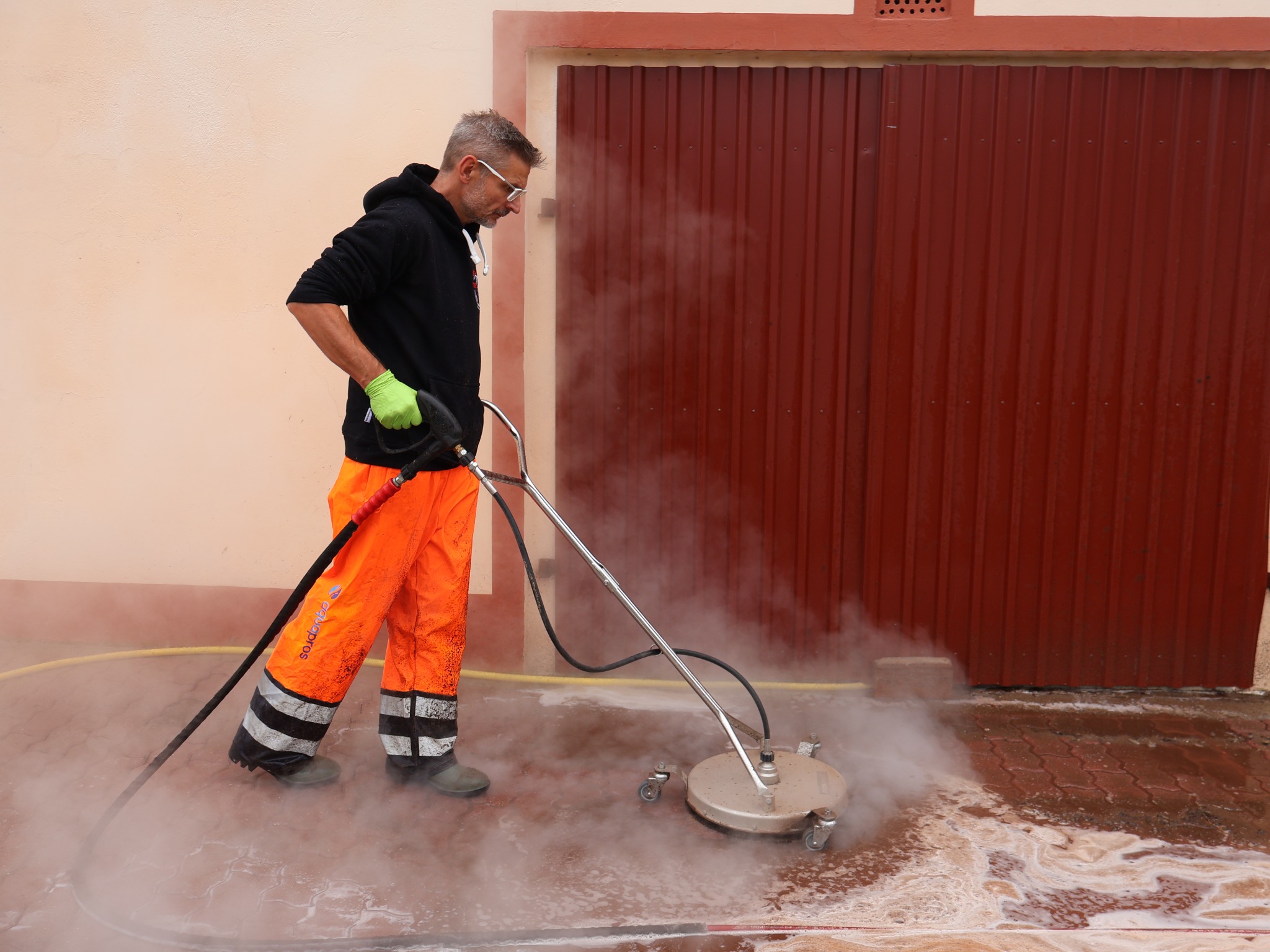 Pracownik w pomarańczowych spodniach roboczych czyści kostkę brukową za pomocą myjki ciśnieniowej z obrotową dyszą, widoczna para wodna i brud spływający po kostce. W tle czerwona brama garażowa.