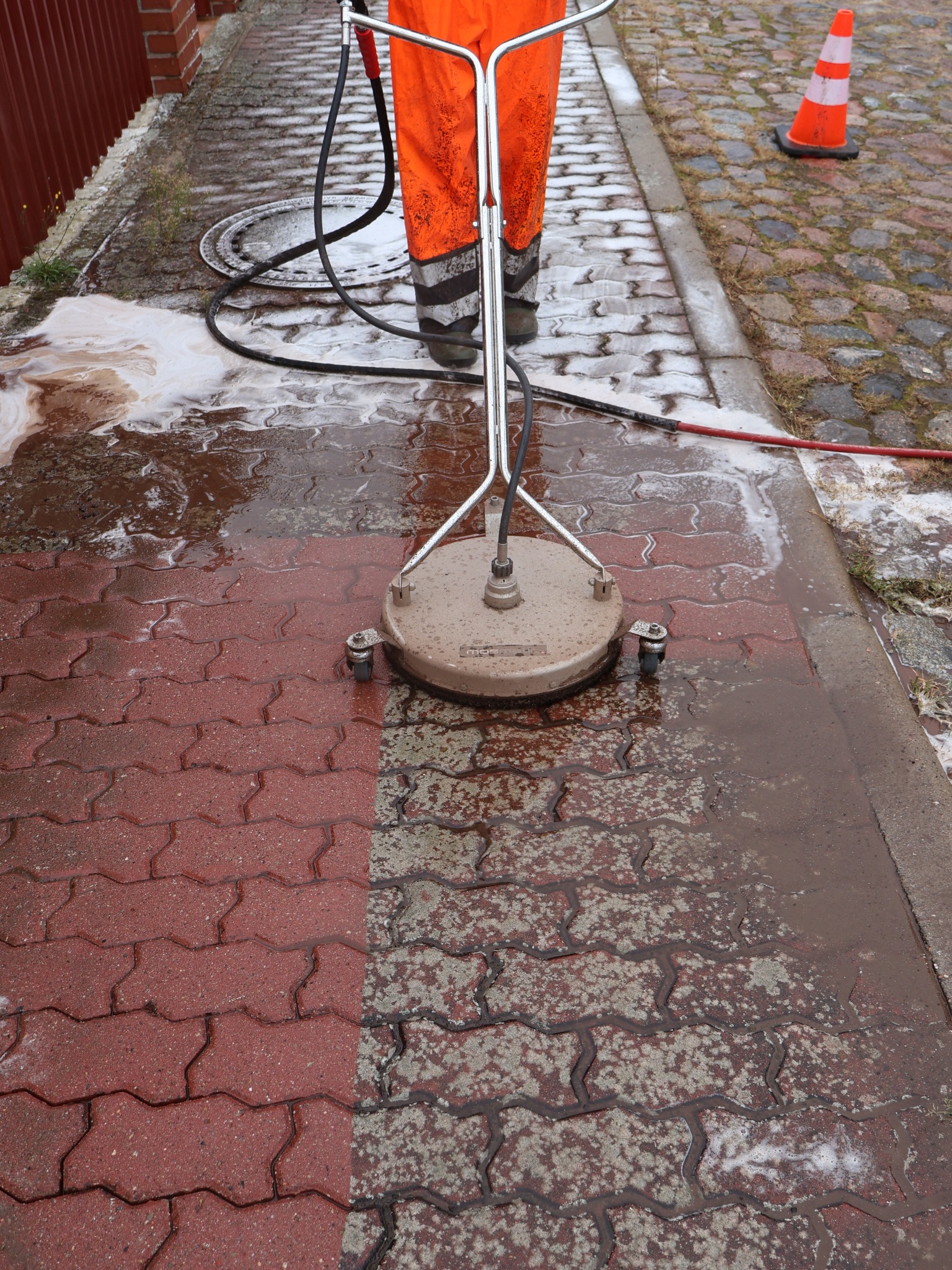 Mycie czerwonej kostki brukowej za pomocą myjki ciśnieniowej z okrągłą dyszą; widoczna wyraźna różnica między czystą a brudną powierzchnią, pracownik w pomarańczowym kombinezonie i pachołek...