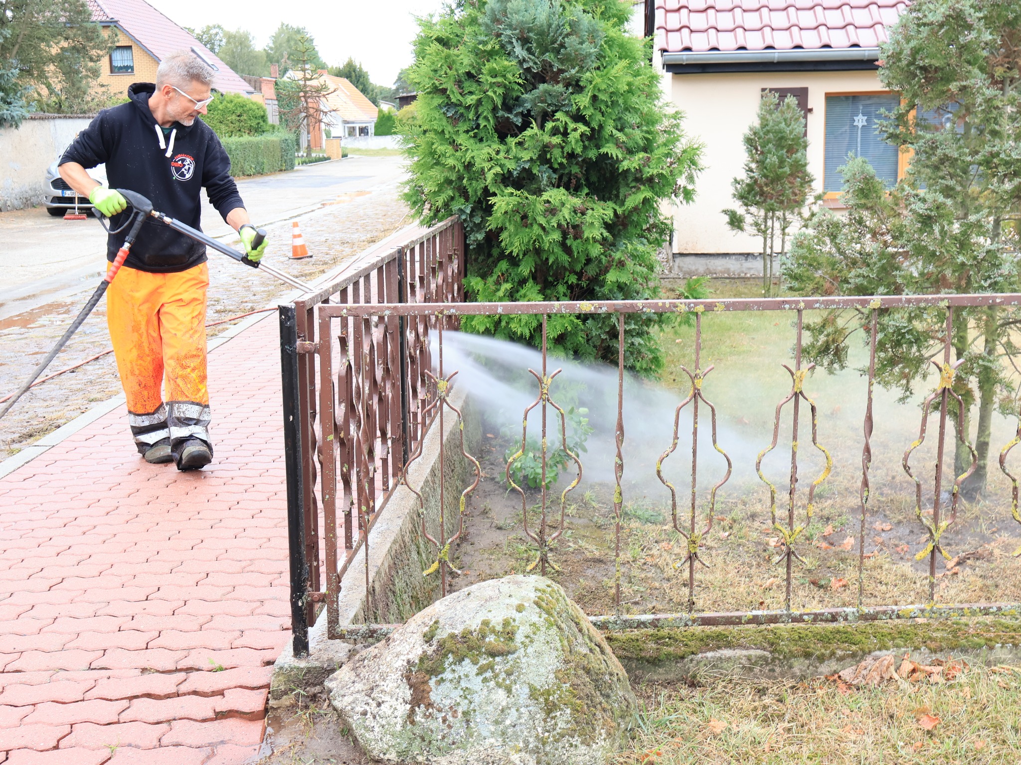 Pracownik w pomarańczowych spodniach i czarnej bluzie z kapturem czyści metalowe ogrodzenie z mchu za pomocą myjki ciśnieniowej. Czerwona kostka brukowa i trawnik w tle.