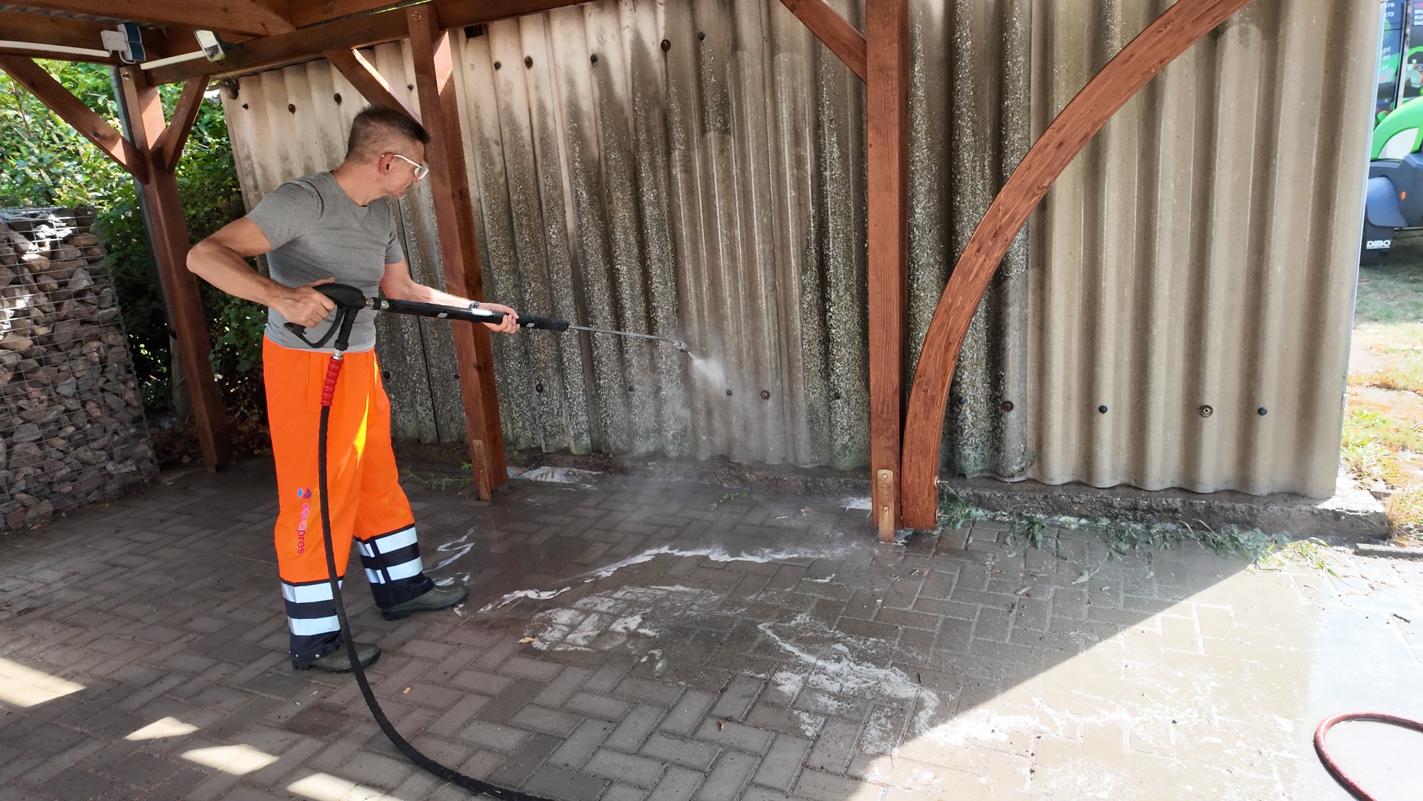 Pracownik w okularach i pomarańczowych spodniach roboczych czyści elewację z blachy falistej myjką ciśnieniową, woda spływa po kostce brukowej.