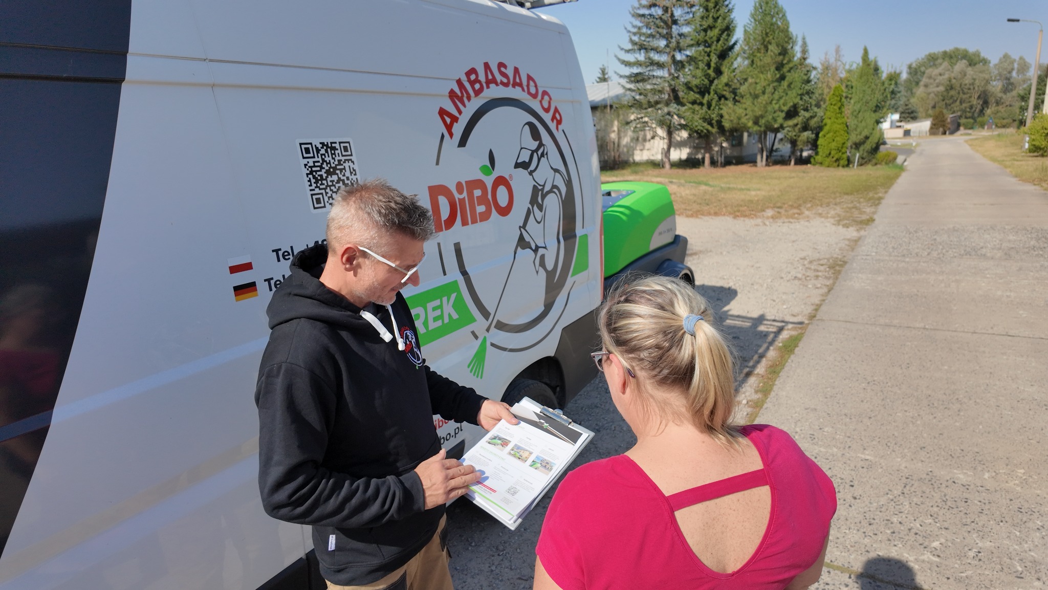 Mężczyzna w czarnej bluzie prezentuje kobiecie folder reklamowy na tle firmowego samochodu z logo Dibo i myjką ciśnieniową.