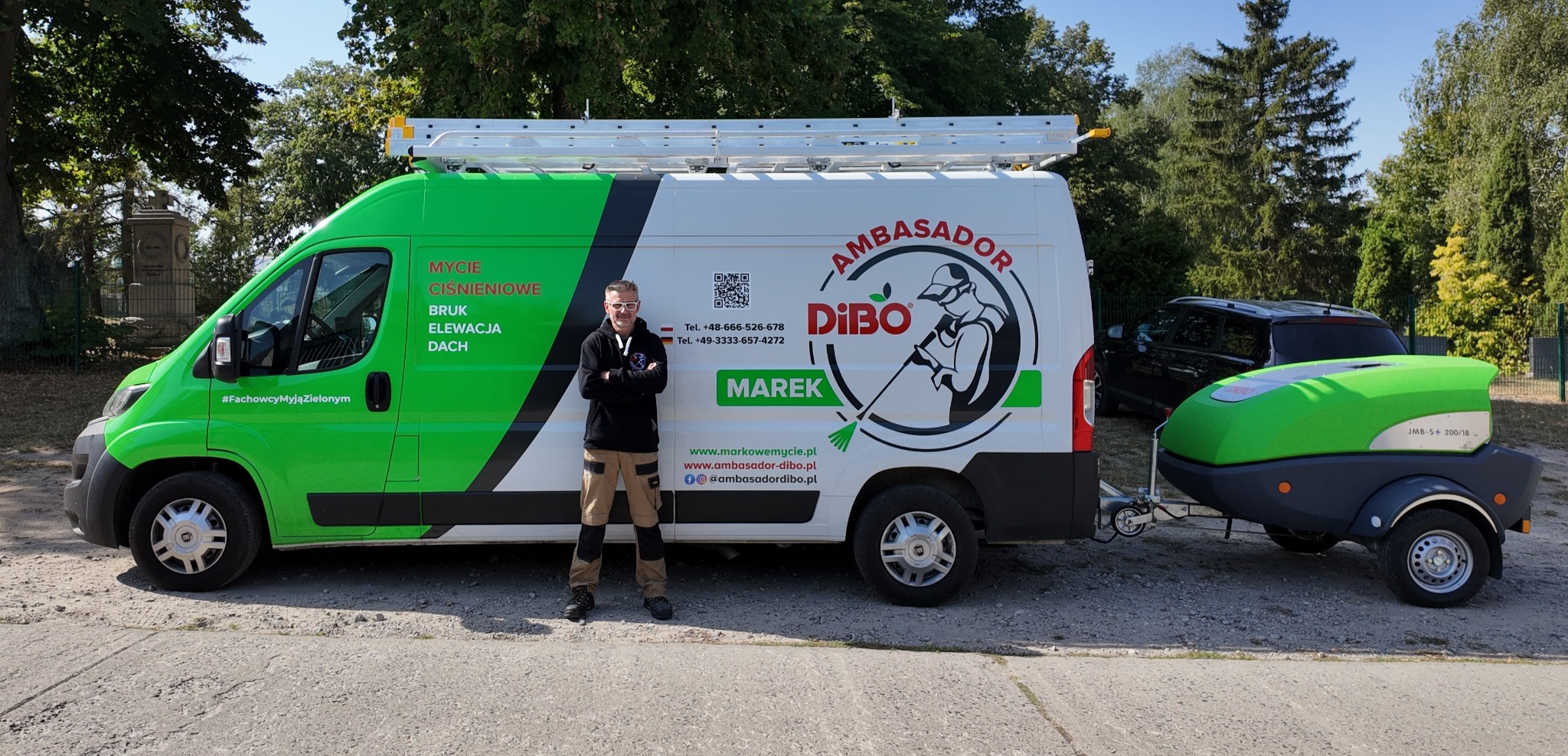 Mężczyzna w okularach, ubrany w czarną bluzę i beżowe spodnie robocze, stoi na tle firmowego, zielono-białego busa z napisem 'Mycie ciśnieniowe BRUK Elewacja Dach' oraz logotypem firmy i przyczepą...