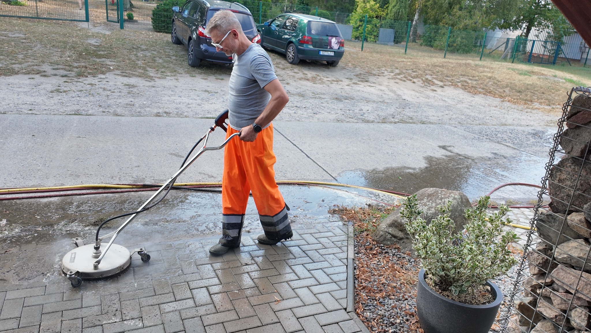 Mężczyzna w pomarańczowych spodniach roboczych czyści kostkę brukową za pomocą profesjonalnej myjki ciśnieniowej; widoczny fragment wyczyszczonej powierzchni, a w tle zaparkowane samochody.