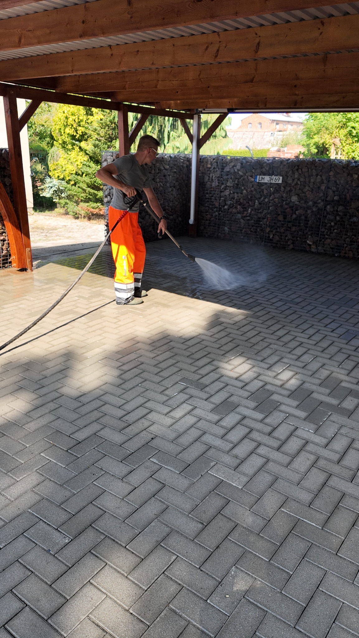 Pracownik w pomarańczowych spodniach roboczych czyści szarą kostkę brukową pod zadaszeniem, używając myjki ciśnieniowej; widoczna ściana z kamieni w koszach gabionowych w tle.