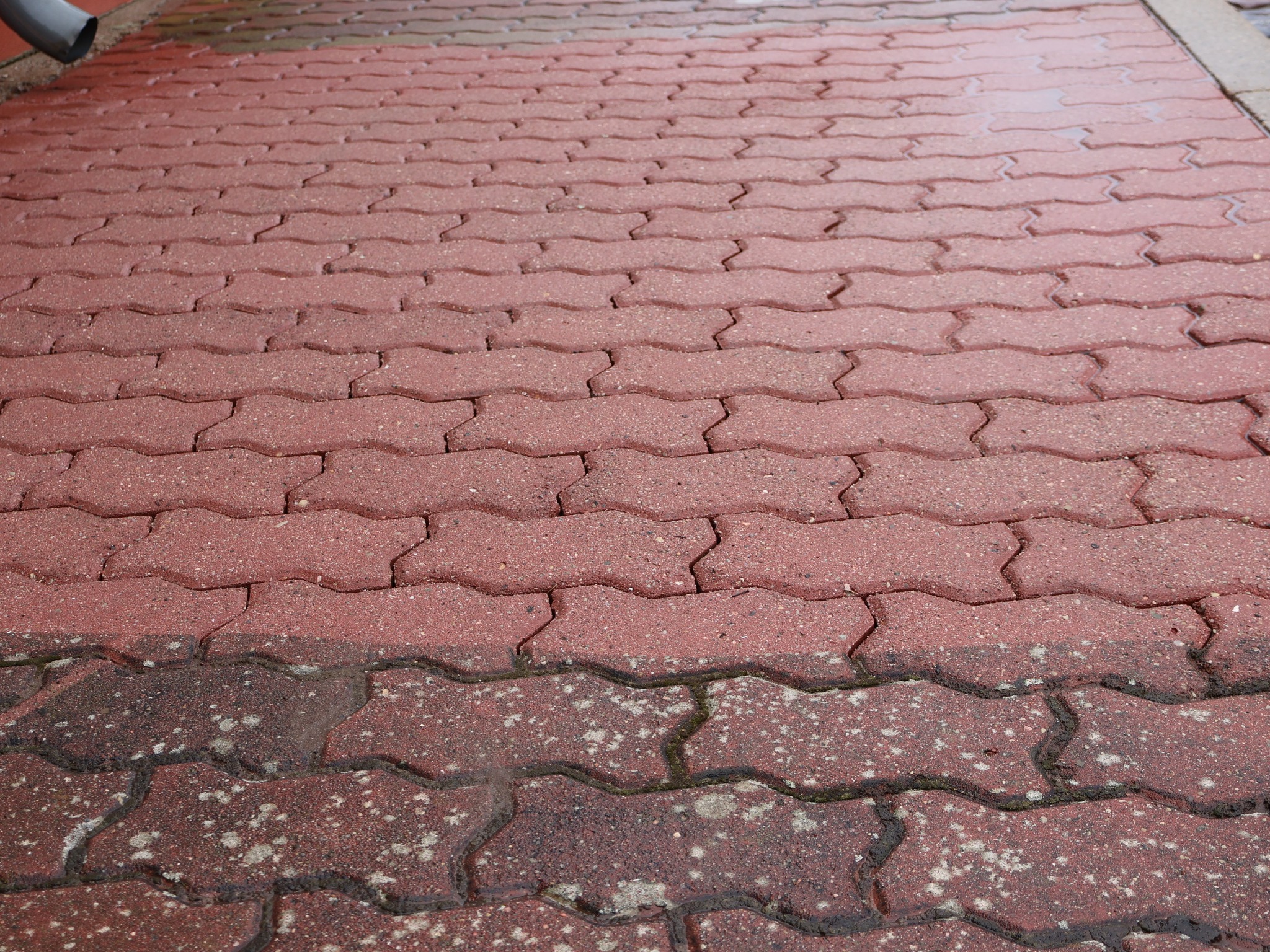 Czerwona kostka brukowa w kształcie zygzaka, częściowo umyta, z widoczną różnicą w kolorze i czystości, z rynną spustową w lewym górnym rogu.